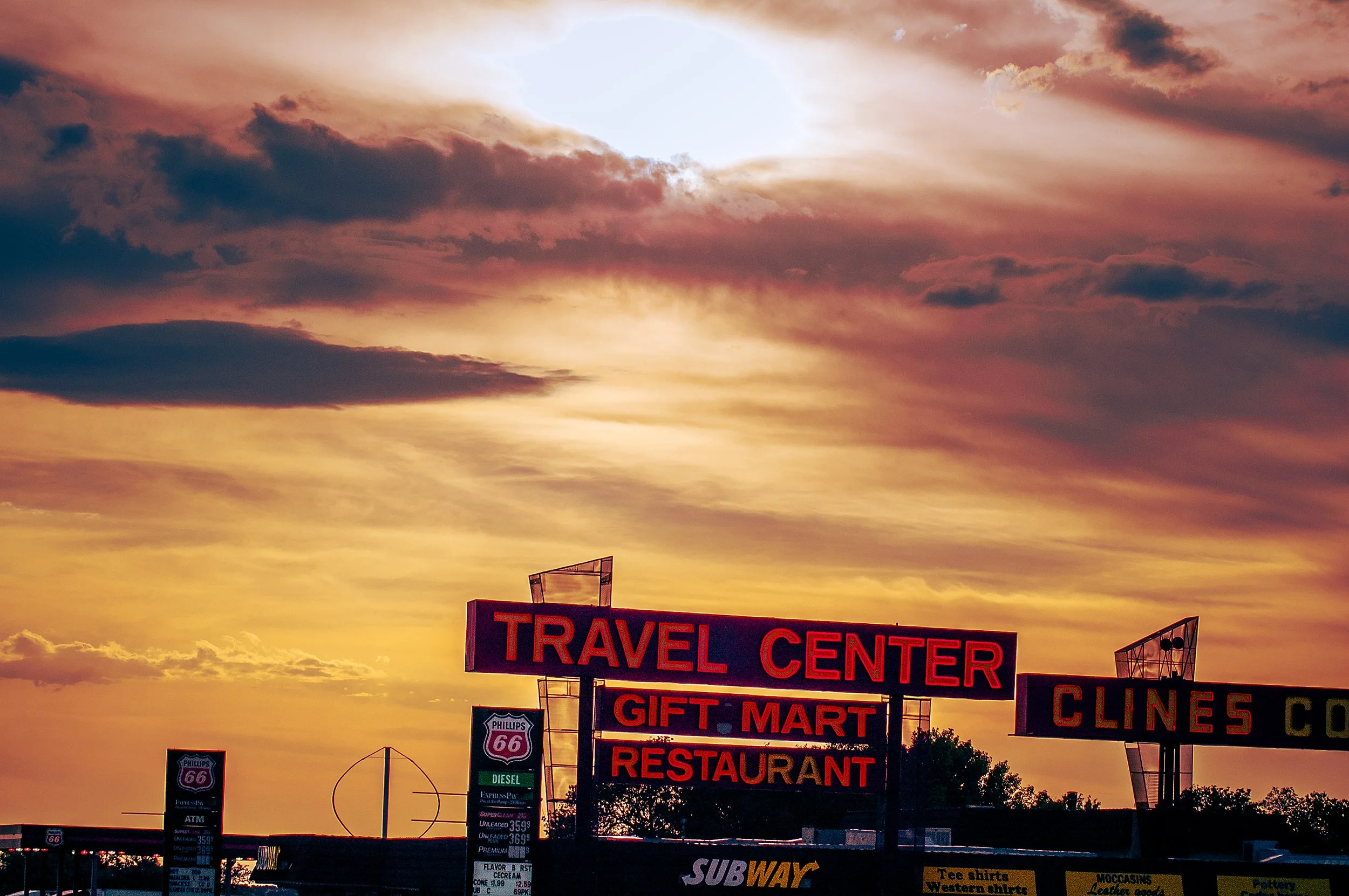 A sky with a mix of orange, pink, and purple clouds during sunset. Below, signs for a travel center, gift mart, restaurant, and a Subway restaurant are visible, along with gas station signs for Phillips 66 and fuel prices.