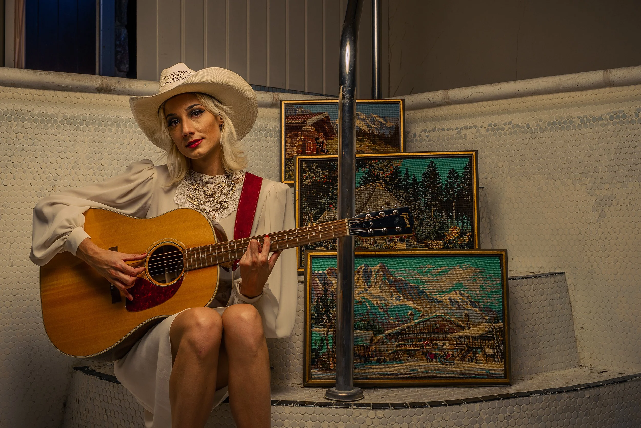 A woman with blonde hair, wearing a large white hat and beige dress, sitting on a bench, playing an acoustic guitar. Behind her are several framed landscape cross-stitch artworks, and the area has a tiled wall with small white hexagons.