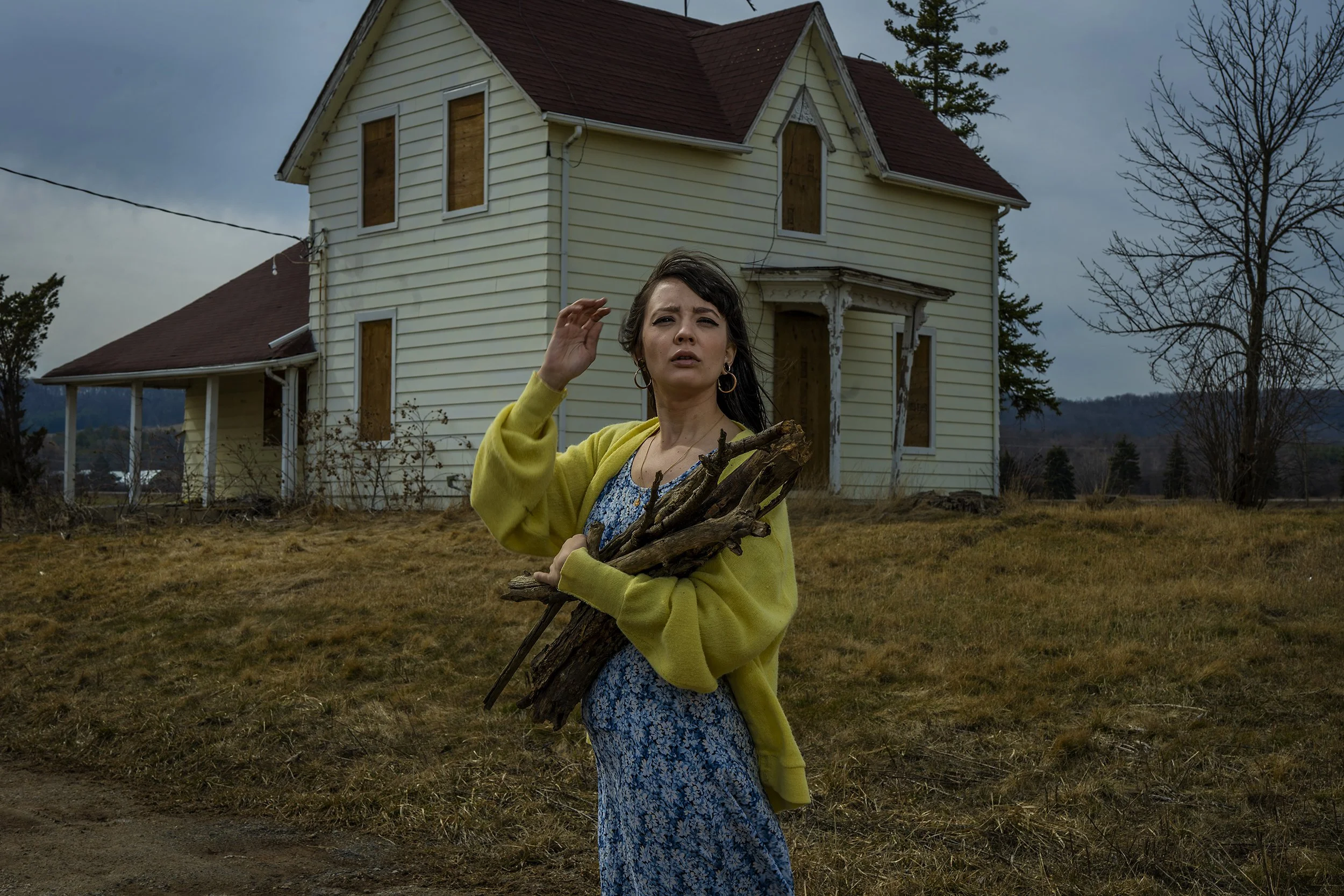 A woman with dark hair wearing a yellow cardigan and holding sticks stands in front of an abandoned, boarded-up house on a cloudy day. The grass is brown, and a leafless tree is visible to the right.
