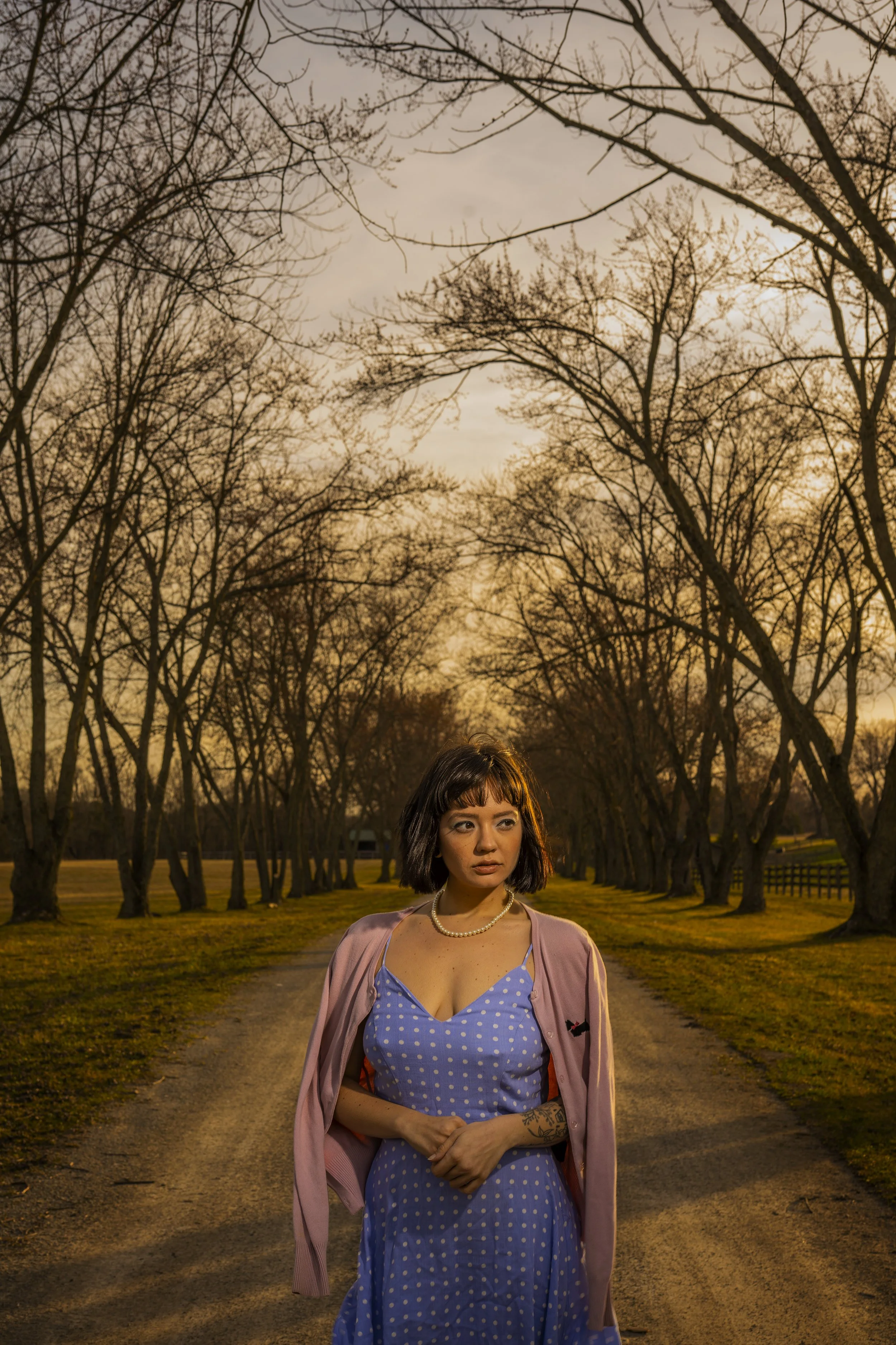 A young woman with shoulder-length dark hair and tattoos on her arm, wearing a blue polka dot dress and pink cardigan, standing on a tree-lined path during sunset.