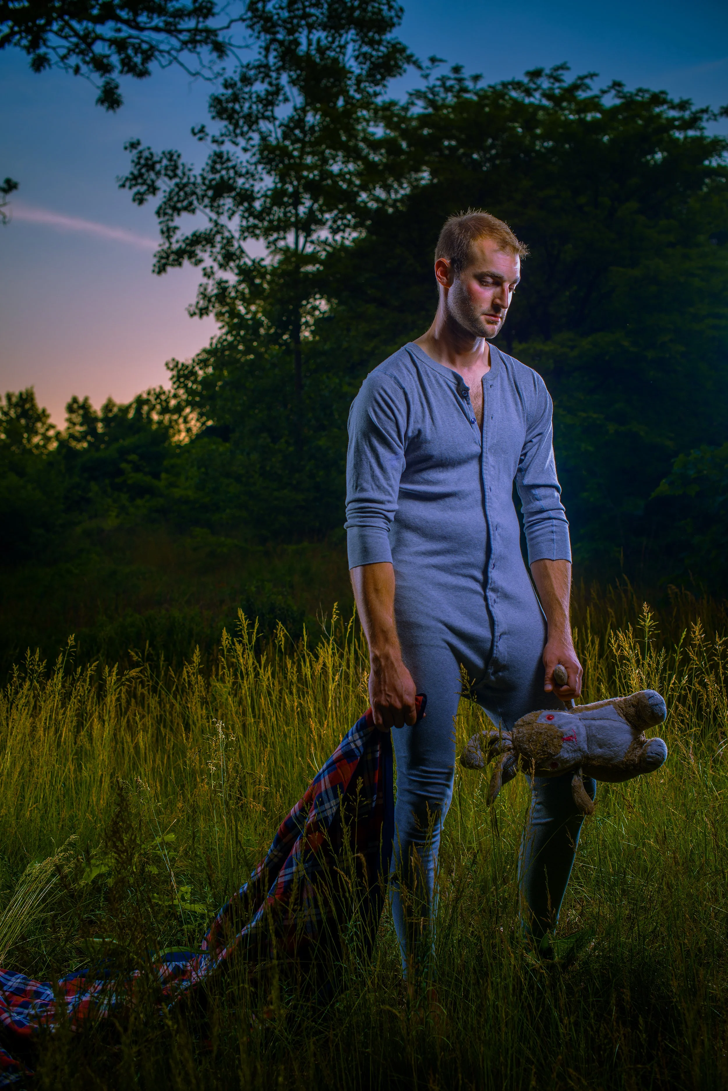 A man standing in a grassy field during sunset, holding a worn stuffed animal and a plaid shirt, appearing somber.