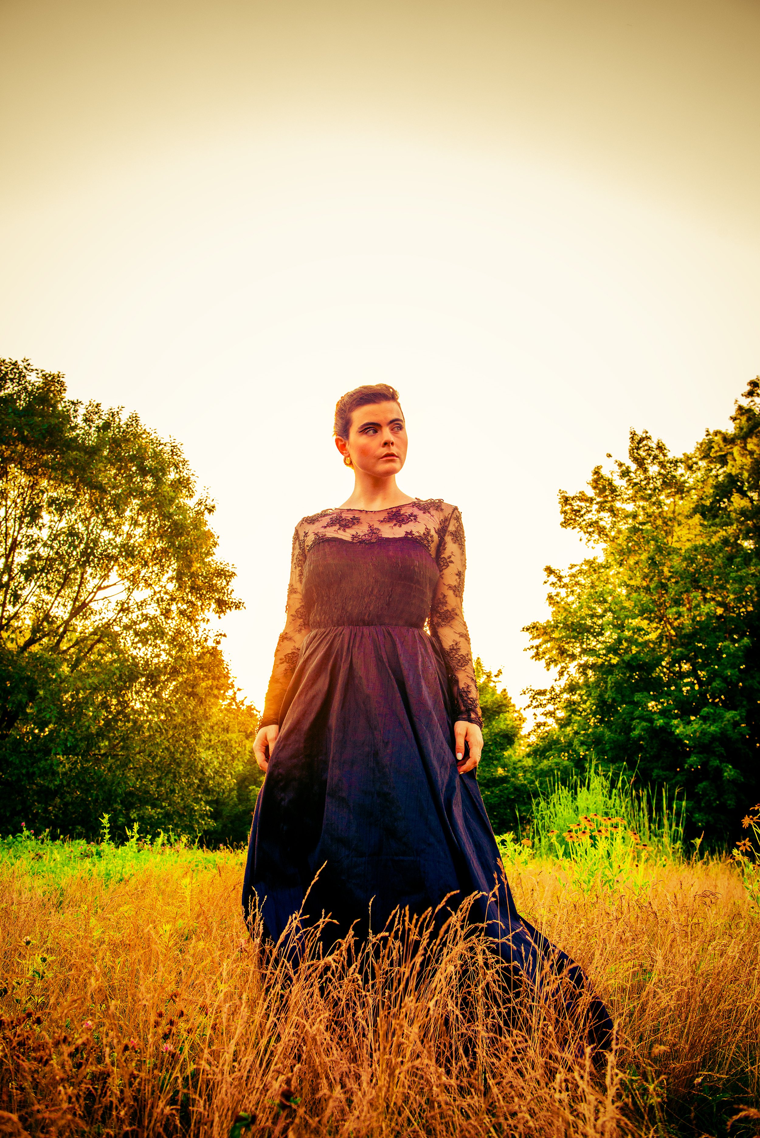 A woman in a long black dress with lace sleeves standing in a field of tall yellow grass during sunset, with trees in the background.