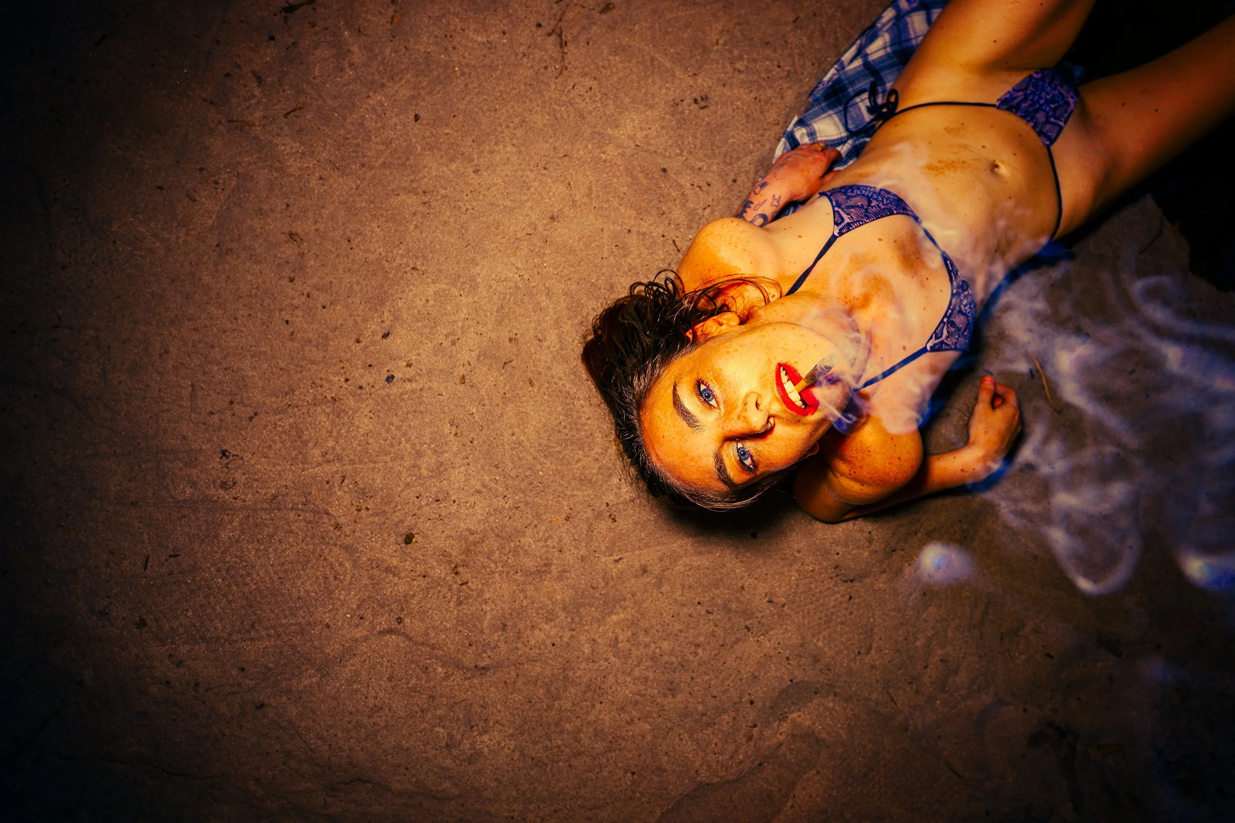 A young woman lying on the ground at night, wearing a blue patterned bikini, with a lit cigarette in her mouth and smoke surrounding her. She has curly hair and is looking upward.