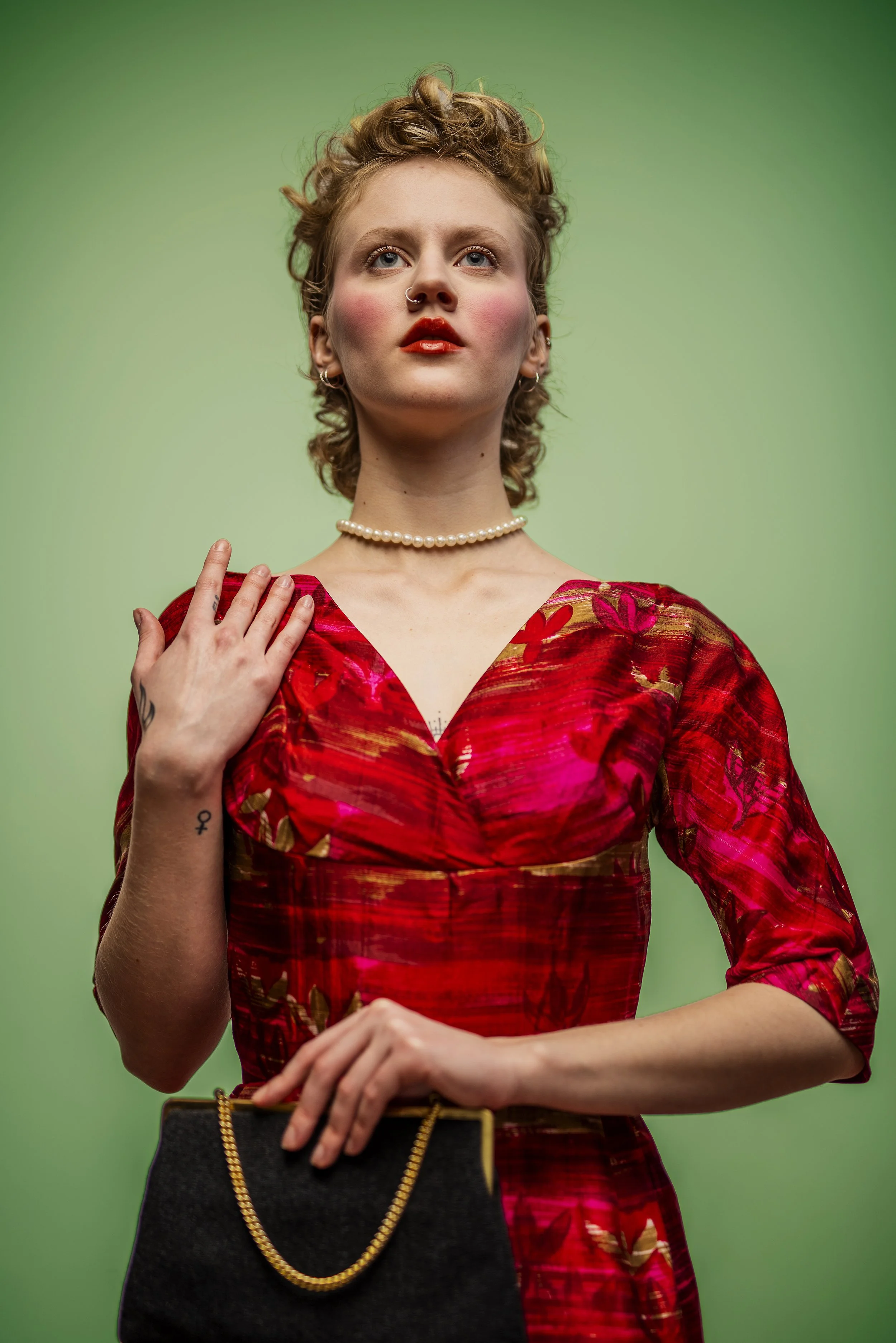 A woman with curly hair wearing a red floral dress, pearl necklace, and lipstick, holding a black handbag with a gold chain in front of a green background.
