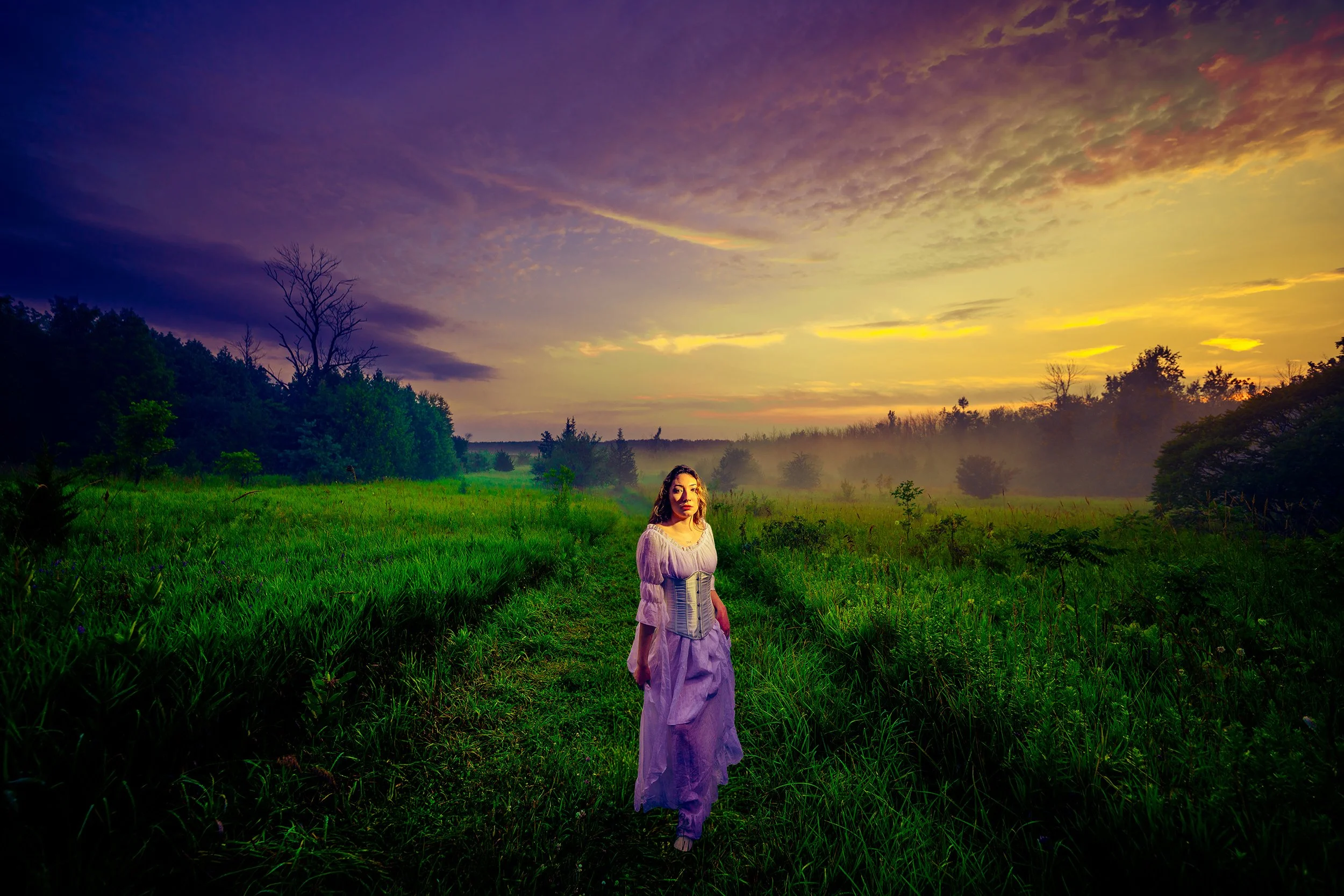 A woman in a purple dress standing in a lush green field during sunset, with a colorful sky filled with clouds and trees in the background.
