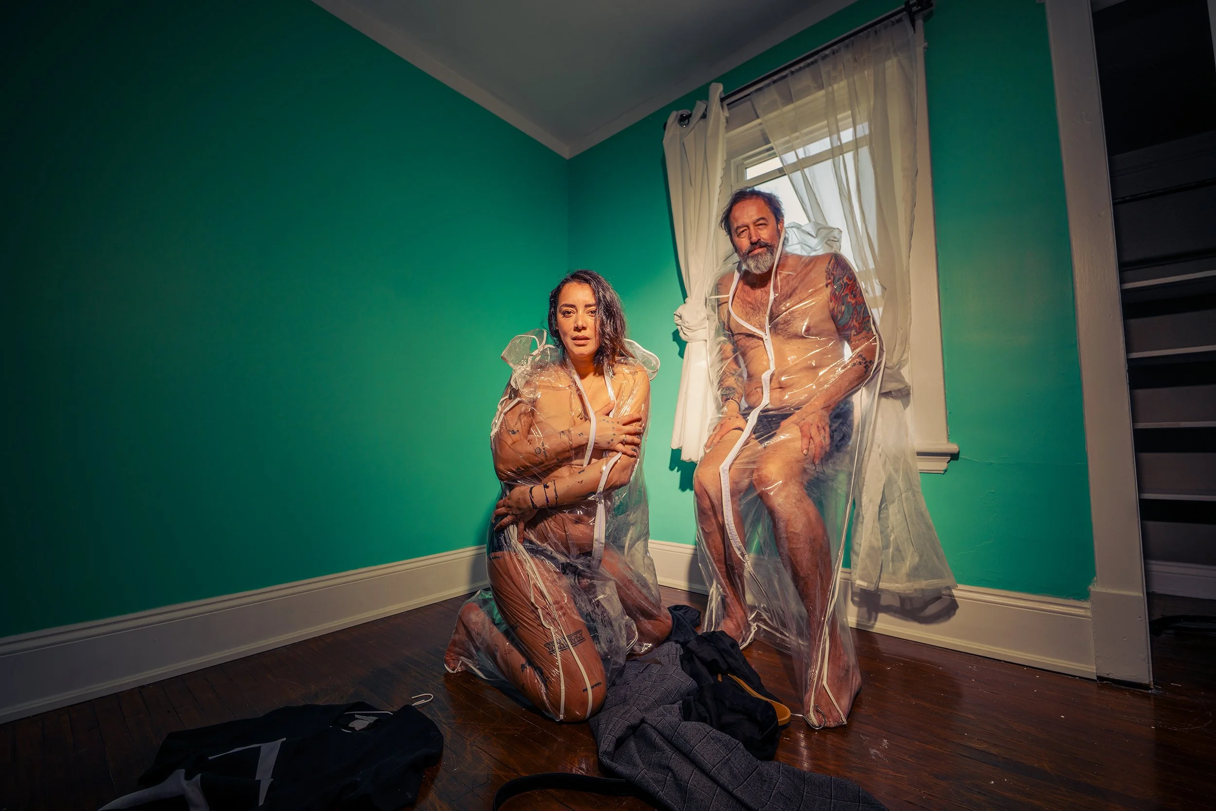 A woman kneeling on the floor and a man sitting on a chair, both dressed in clear plastic suits, in a room with green walls and a window with white curtains.