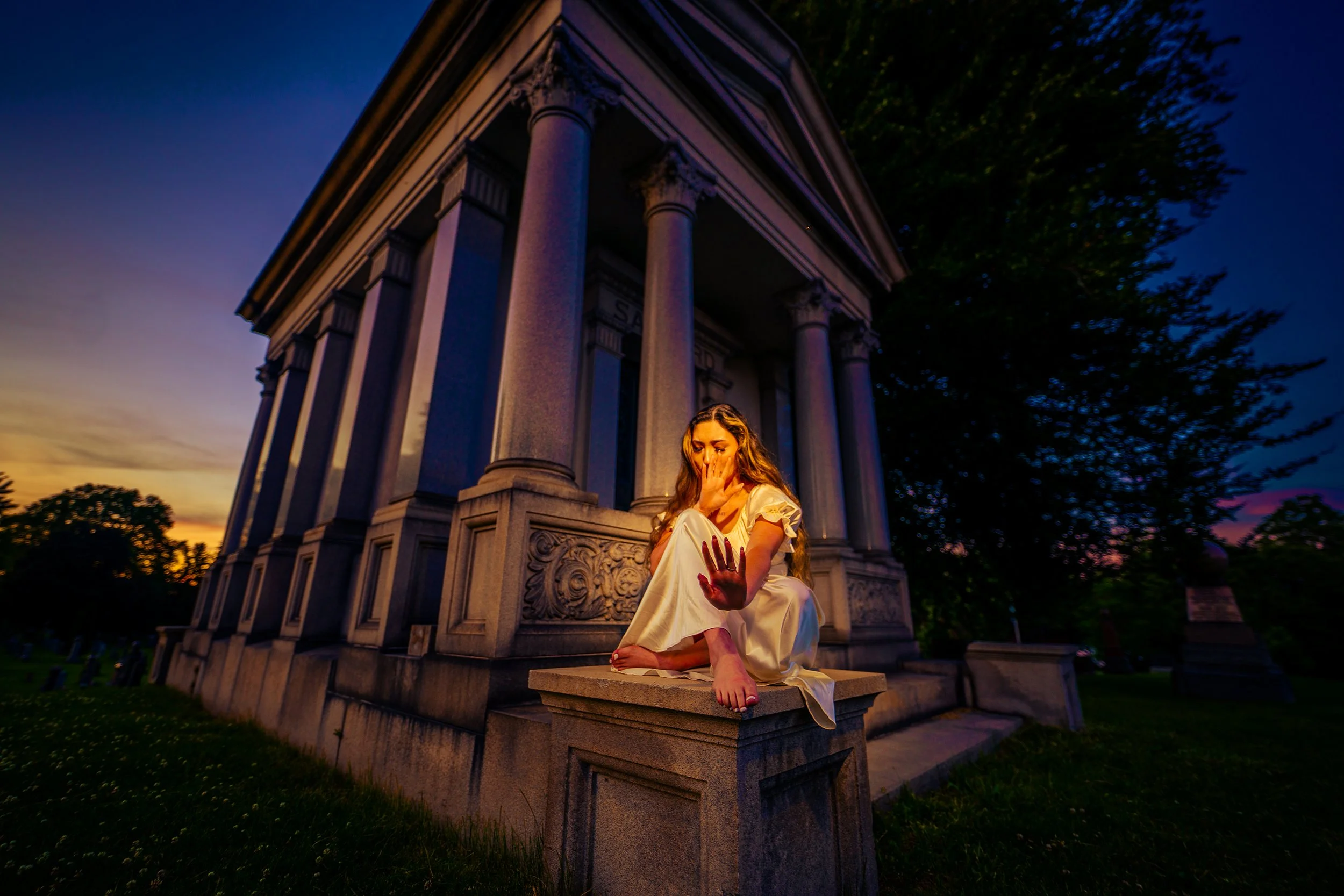 A woman with long hair wearing a white dress sitting on a stone monument in front of a large building with columns at sunset, holding her face and extending her hand forward with red-painted nails.