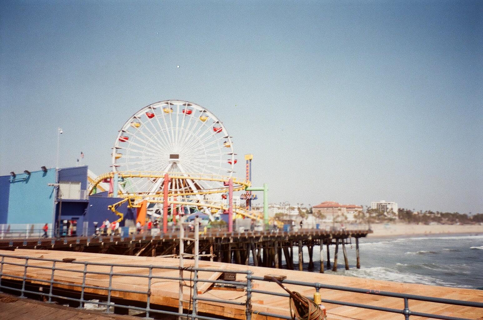 a colorful amusement park on a pier with a large Ferris wheel, roller coaster, and other rides, facing the ocean during daytime.