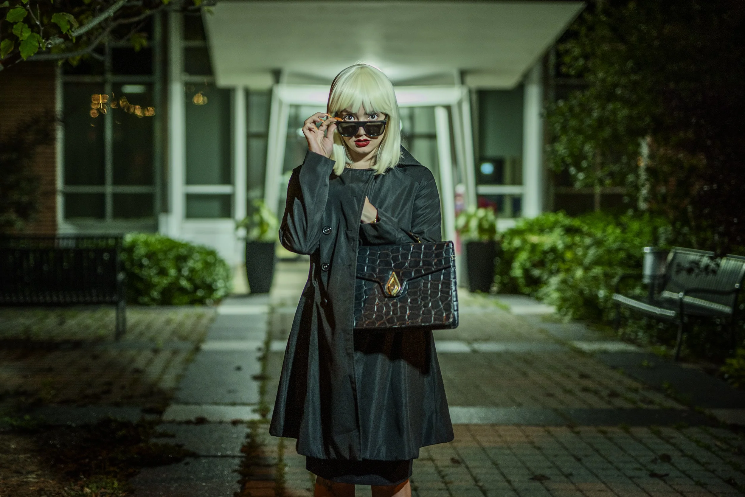 Woman with blonde hair and dark sunglasses standing outside at night, wearing a long black coat and holding a black handbag, with modern building and benches in the background.