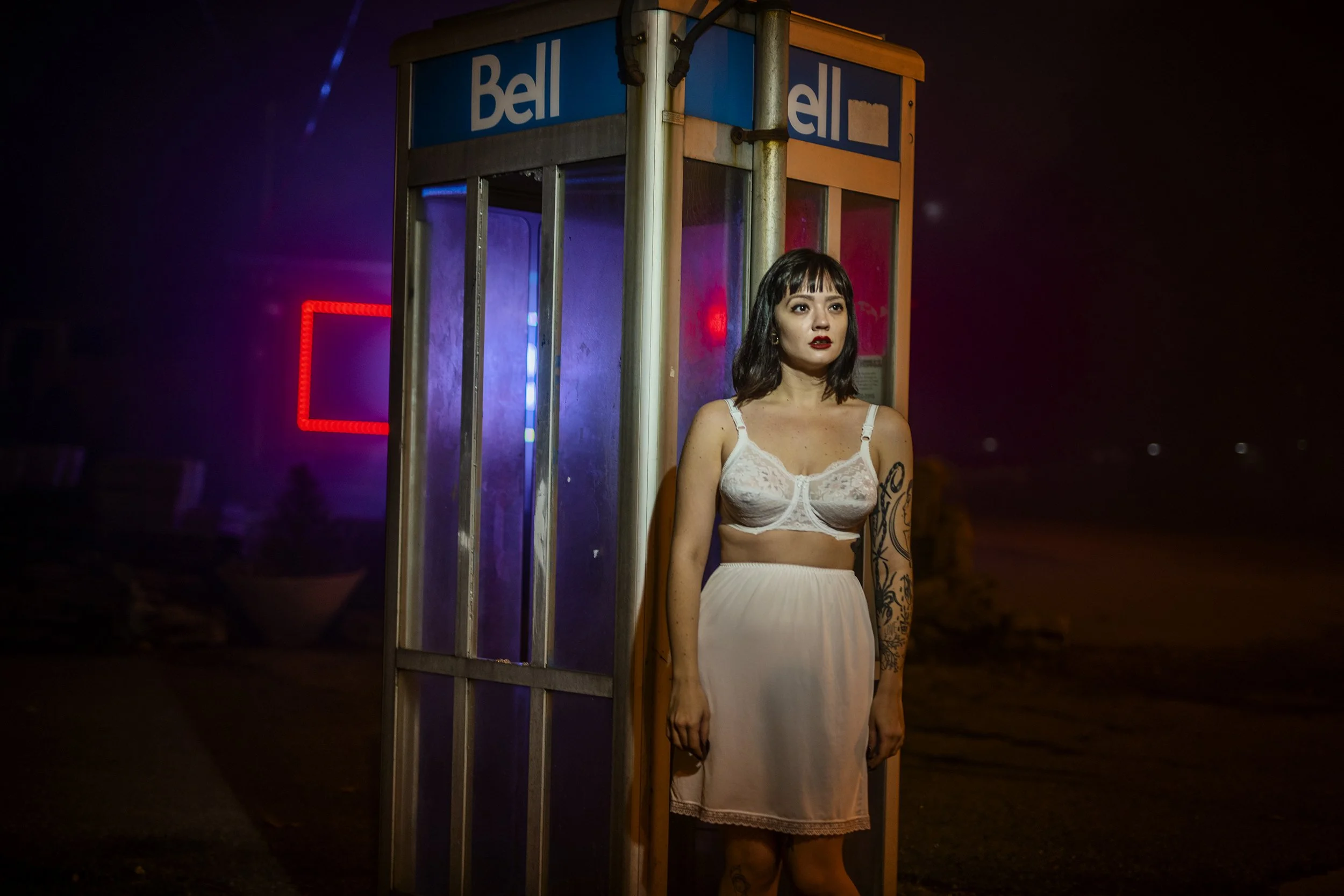A woman with dark hair and tattoos on her arm standing in front of a phone booth with neon lights at night.