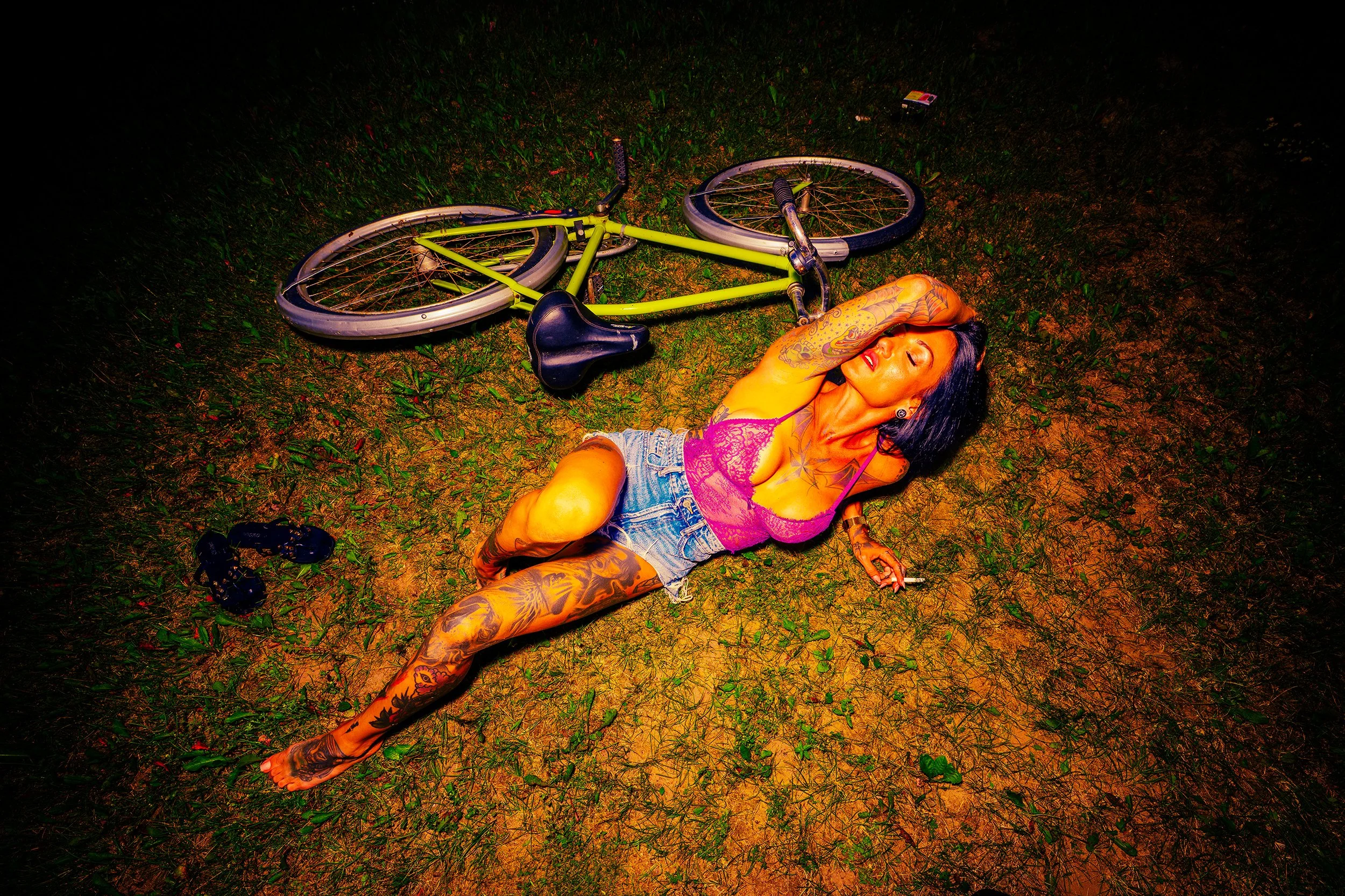 A woman with numerous tattoos lying on the grass at night next to a green bicycle. She has dark hair, is wearing shorts and a pink lace top, and appears to be relaxed or asleep.
