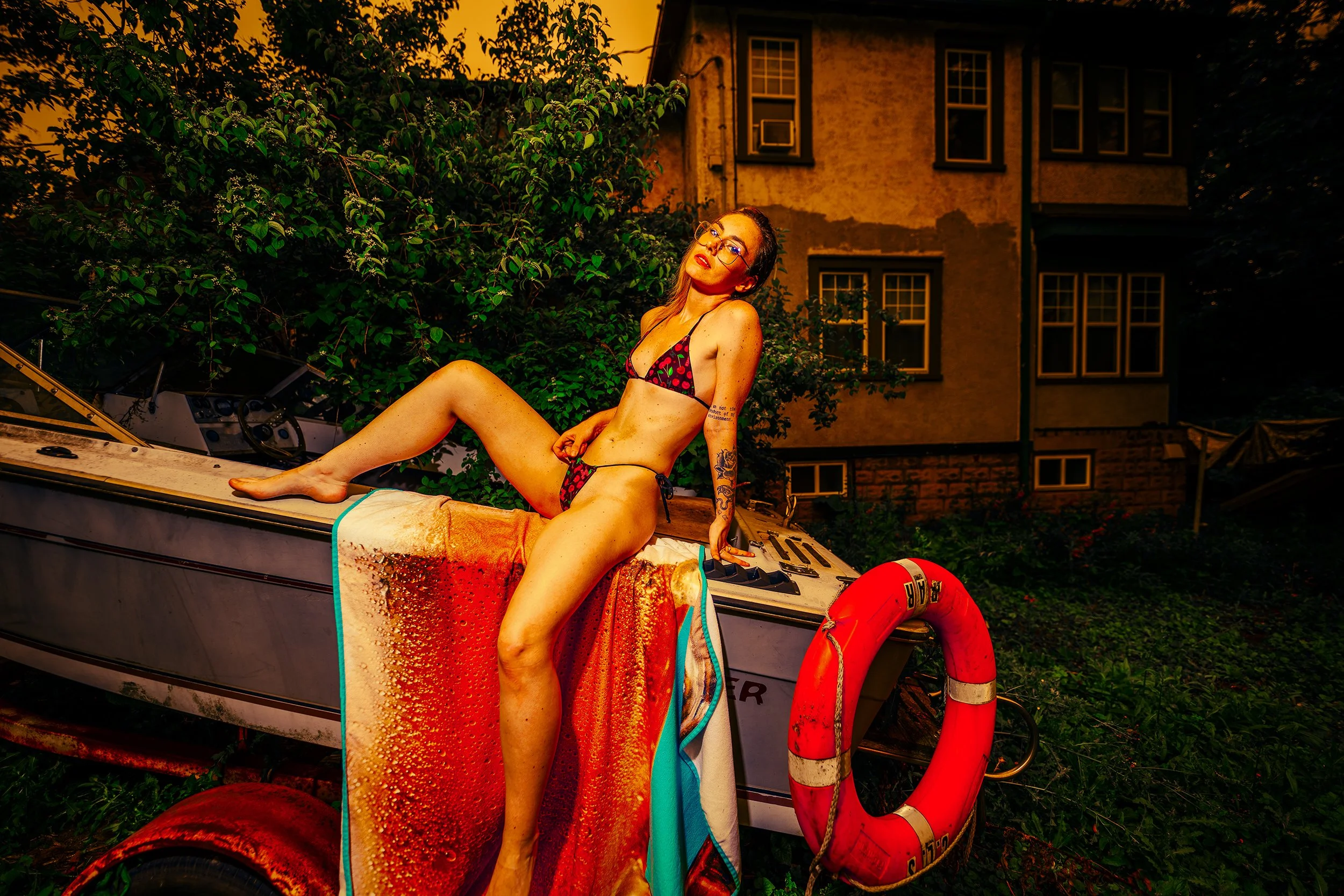 A woman wearing glasses and a bikini with a cherry pattern sitting on a boat at sunset. She has tattoos on her arm and is looking towards the camera. The boat is docking near a house with multiple windows and a yellow-orange sky in the background.