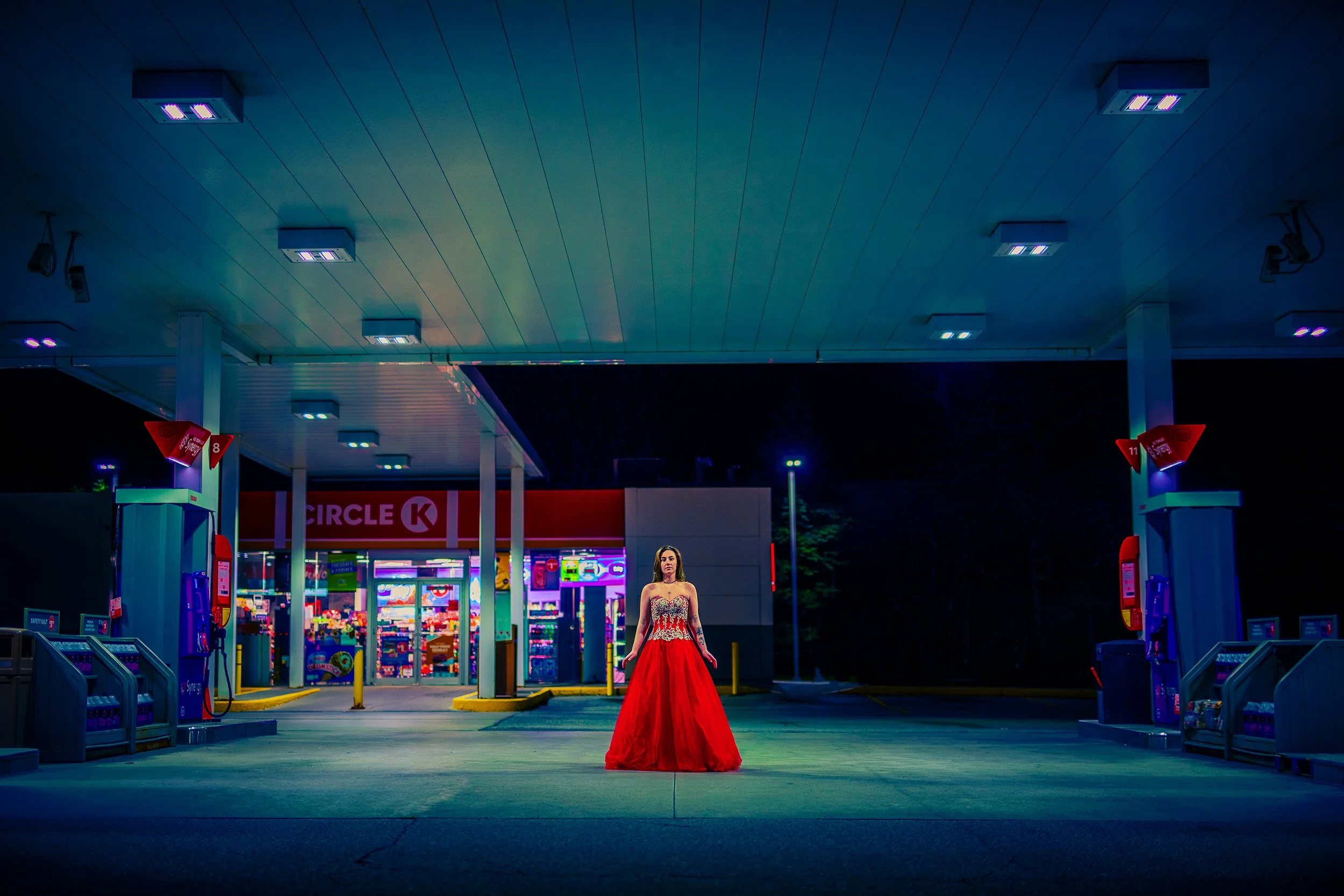 A woman in a red evening gown standing alone in a brightly lit gas station at night, with a colorful convenience store in the background.