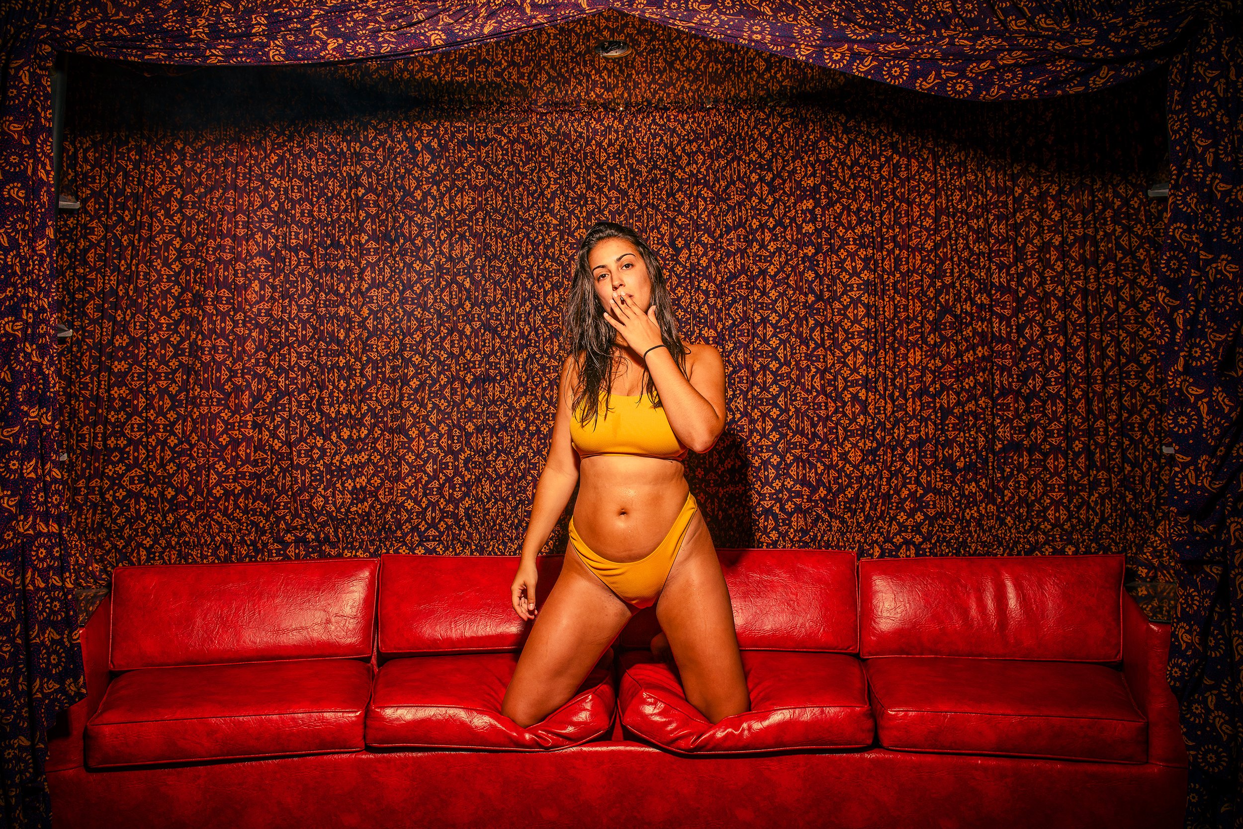 A woman wearing a yellow bra and matching underwear is sitting on a red leather couch in front of a backdrop with intricate orange and dark red patterns on dark fabric.