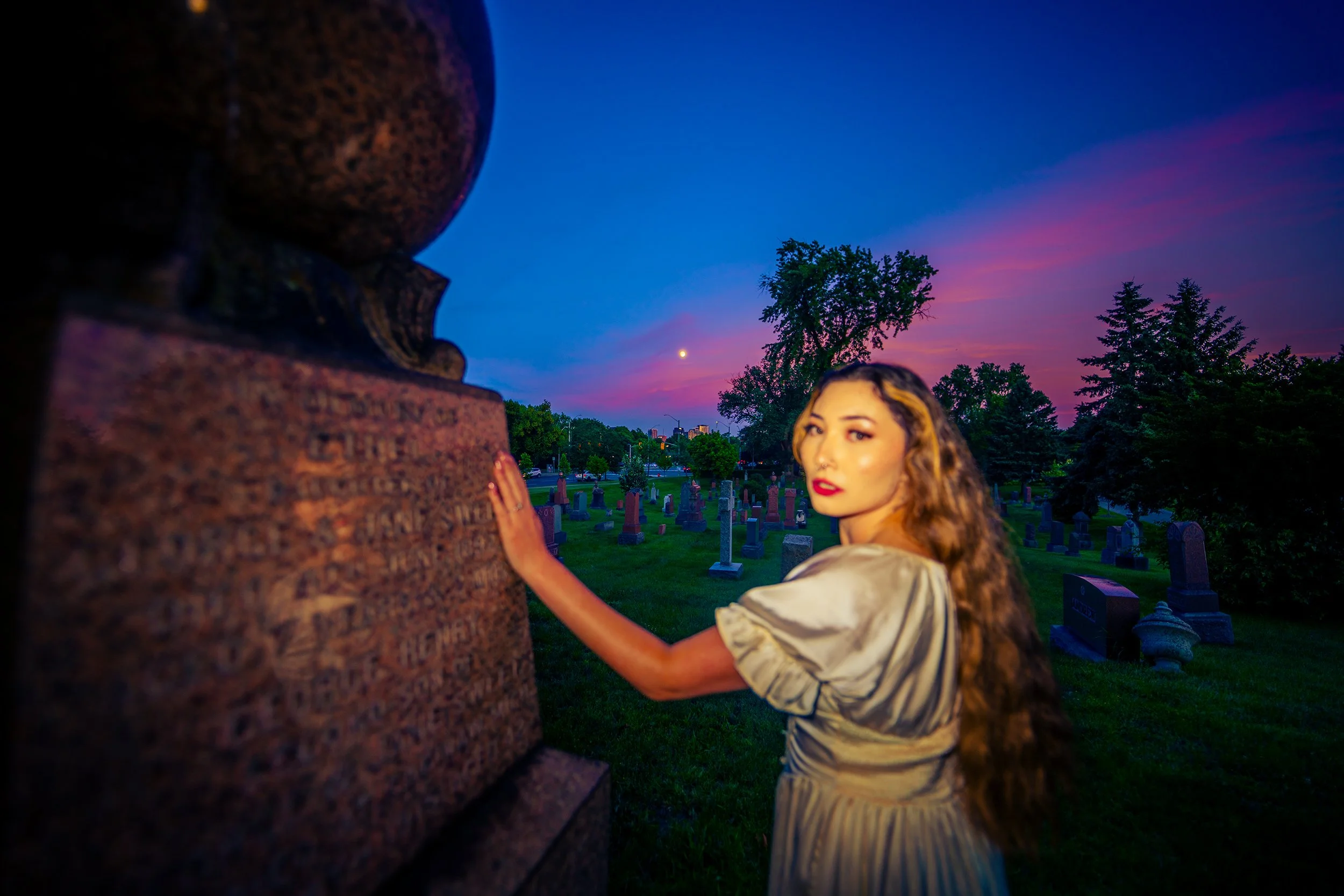 A woman with long, curly hair and wearing a shiny, cream-colored dress stands near a cemetery under a colorful, twilight sky. She is touching a large, upright tombstone with her hand and looking into the distance, with trees and more tombstones visib