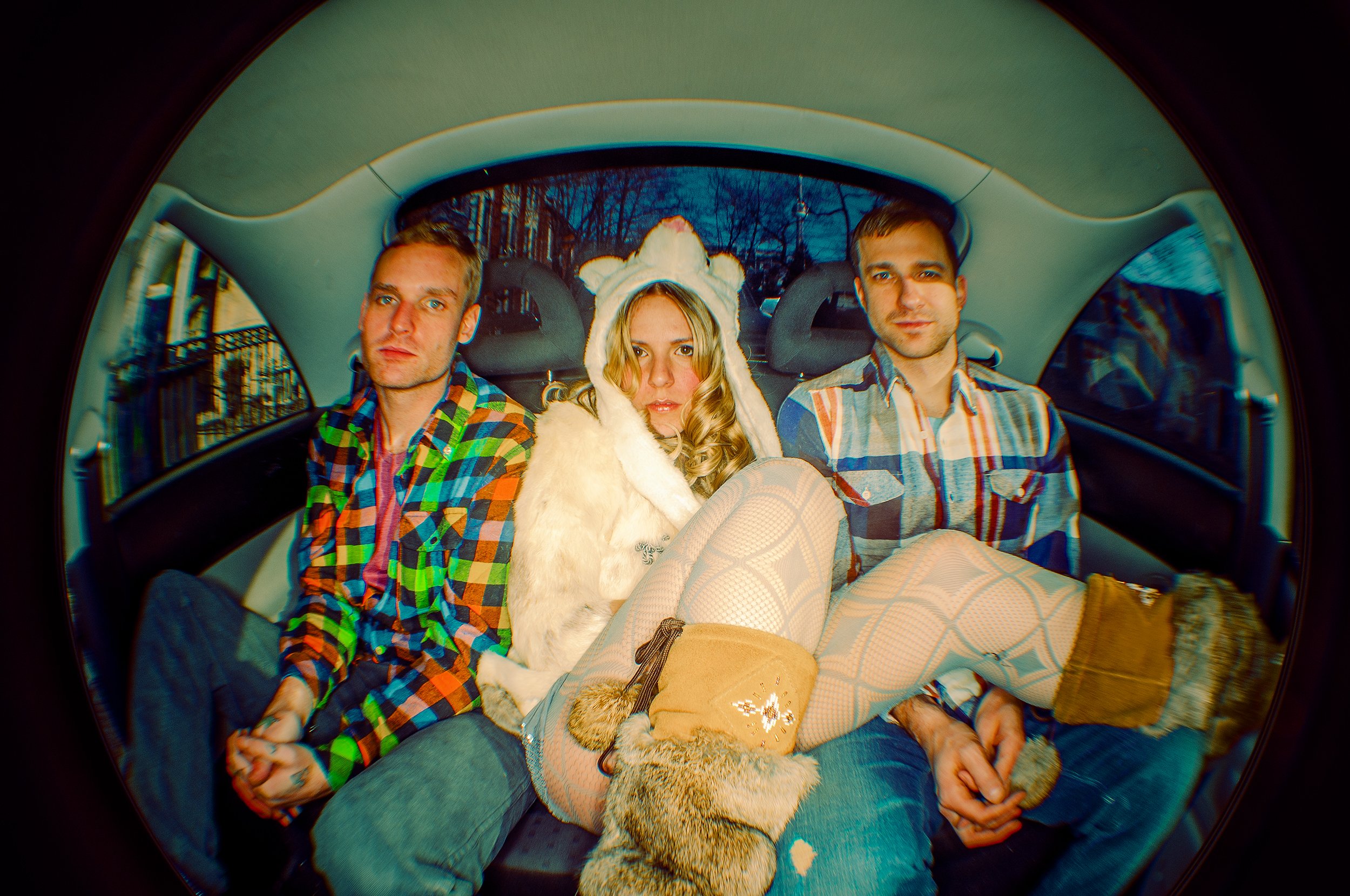 Three people sitting in the back of a car, one woman in the middle wearing a white animal-themed hat, and two men on either side wearing plaid shirts, with fishnet stockings and boots, and a pair of furry shoes visible in the foreground.