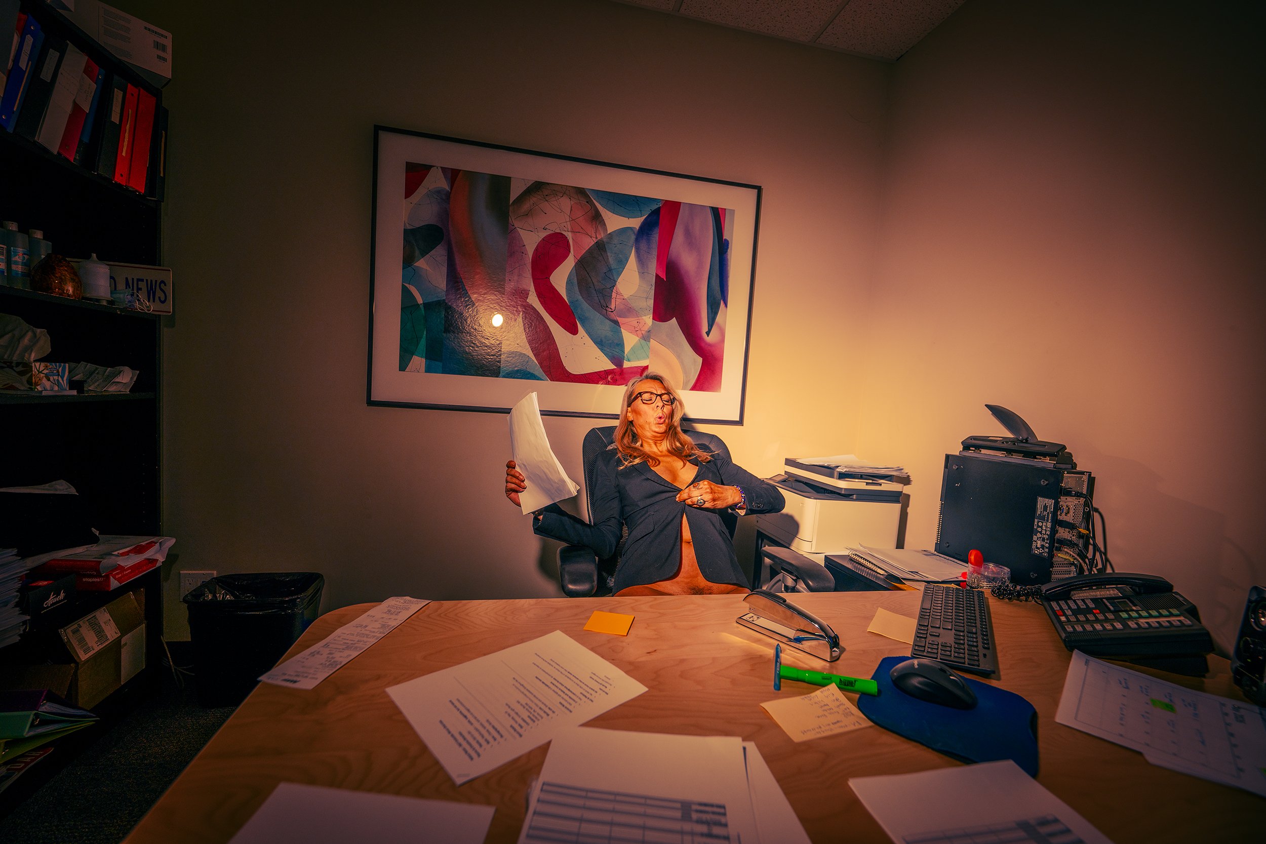 An office with a cluttered desk, scattered papers, and office supplies. A woman with glasses and long hair is sitting in a chair, making an expressive face. The office has a colorful abstract painting on the wall, shelves with binders and items, and 
