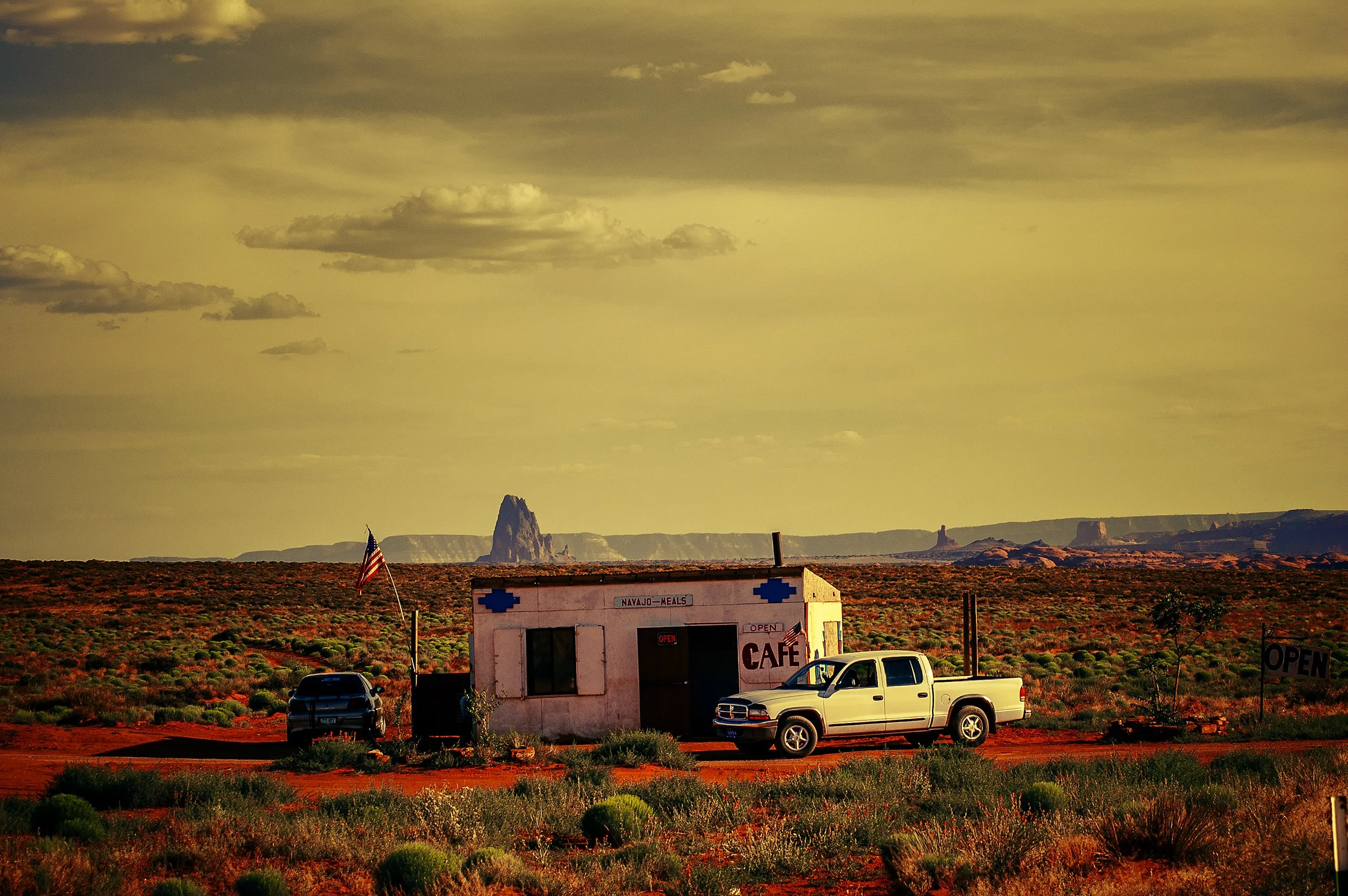 A roadside cafe building with an American flag, parked cars, and a desert landscape with rock formations and a dramatic sky with clouds.