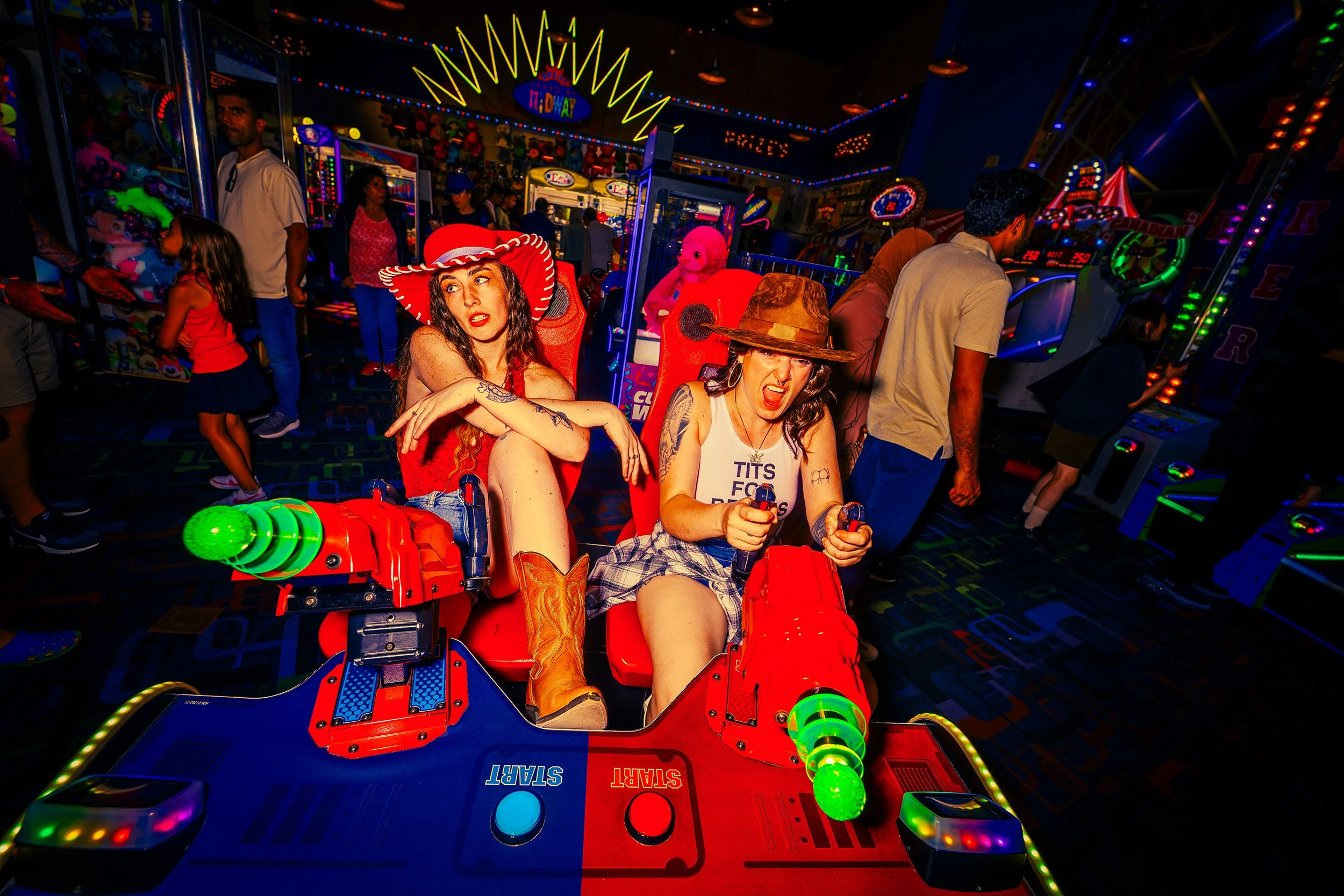 Two women playing an arcade shooting game in a brightly lit amusement park. The game has a red and blue seat with green cannons. The women are focusing intently, one wearing a cowboy hat and a white tank top, the other wearing a large red hat and red