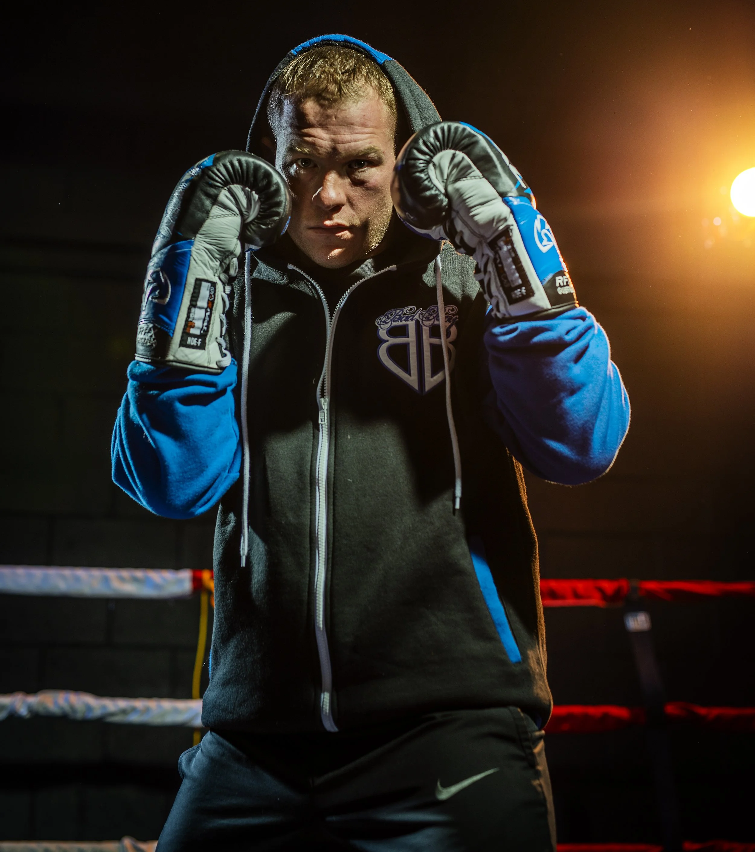 A male boxer in a boxing gym posing with gloves, wearing a hoodie and athletic pants, with a boxing ring in the background and bright lighting.