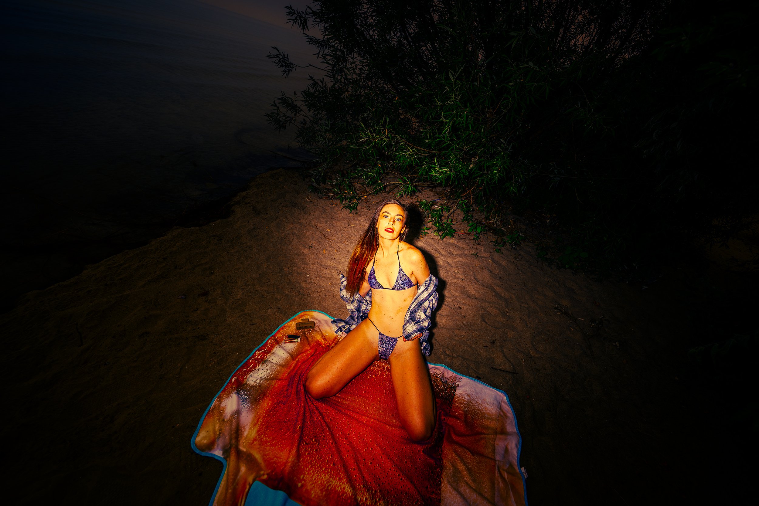 A woman with long hair sitting on a blanket on the beach at night, wearing a bikini and a plaid shirt, surrounded by dark trees.