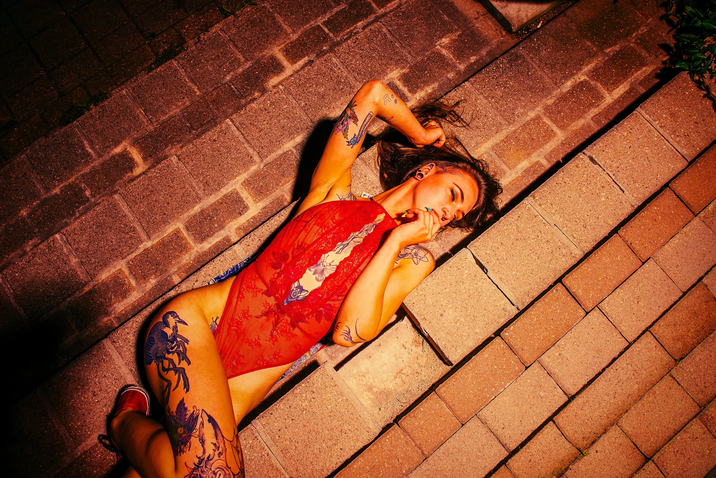 A woman with multiple tattoos lying on a brick sidewalk at night, wearing a red lace dress and red shoes, with her eyes closed and hair spread out.
