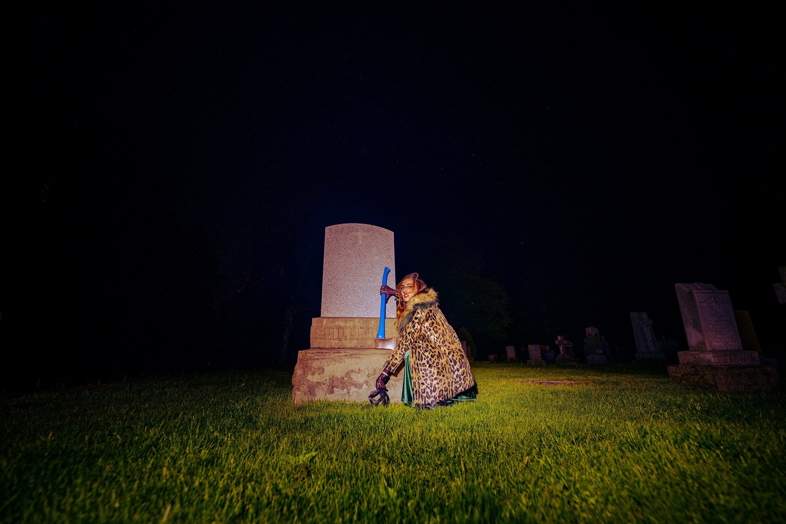 Nighttime photo of a woman standing on grass in front of a large tombstone, holding an axe, wearing a leopard print coat, with head tilted and looking at the camera.