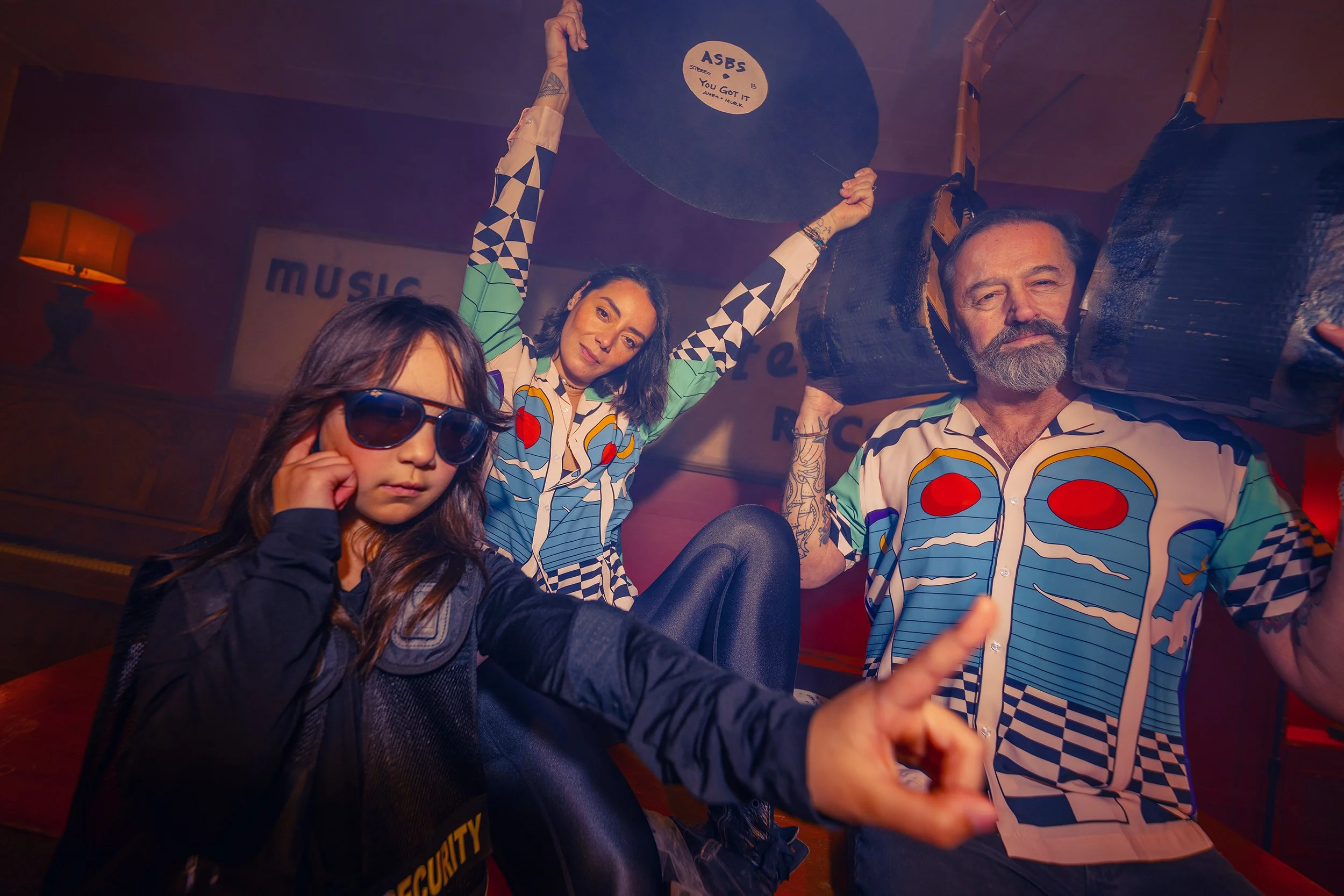 Three people in colorful, patterned clothing in a dimly lit room with a sign that reads "Music" in the background. The girl in the foreground wears sunglasses and a black jacket, pointing with her right hand. The woman in the middle holds a vinyl rec