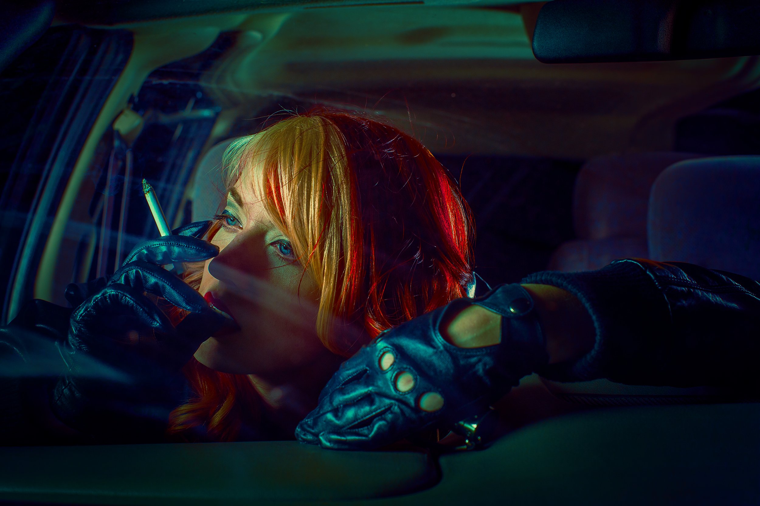 A person with vibrant red hair and blue eyes, wearing black leather gloves, is seated inside a vehicle with a dark interior. The lighting creates neon-like reflections on their hair and gloves.