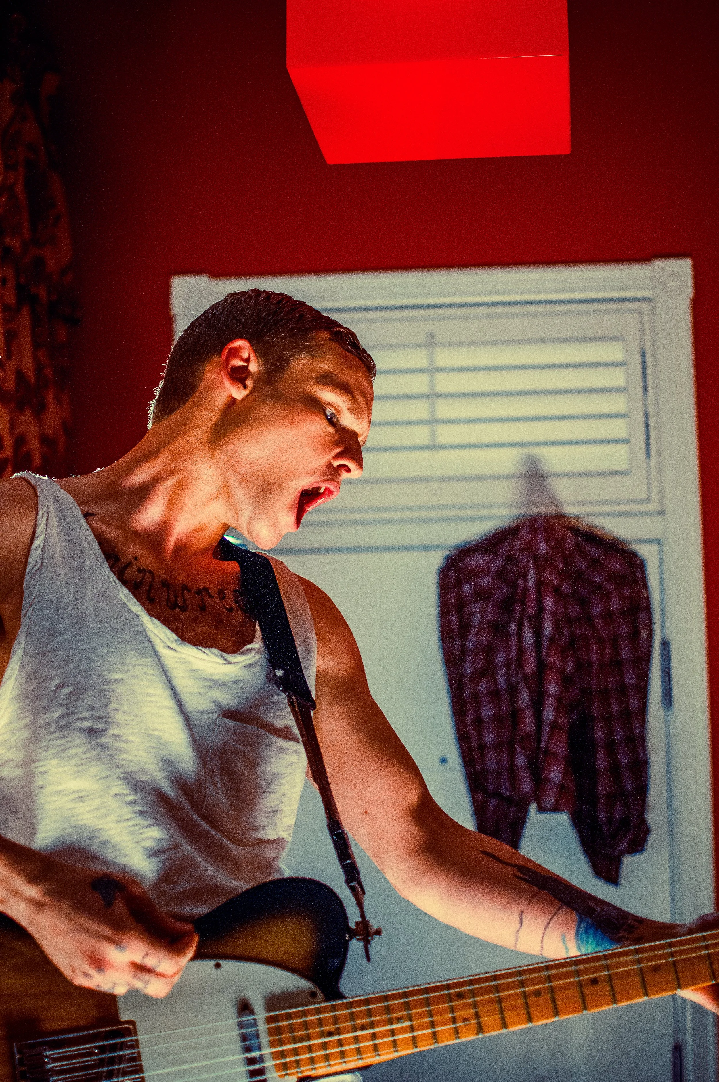Person with short dark hair and tattoos on arms playing an electric guitar, wearing a sleeveless shirt, in a room with a window and a red square ceiling light.