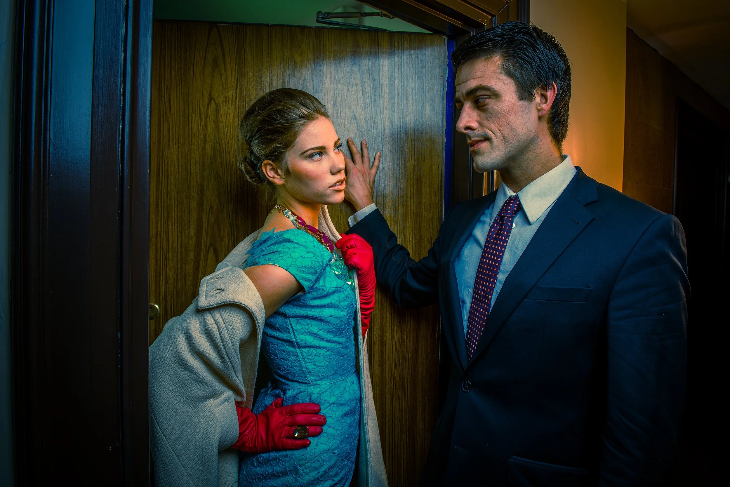 A woman in a blue dress and beige coat confronts a man in a dark suit in an elevator, gazing intensely, with the man gently touching her face as they have a tense moment.