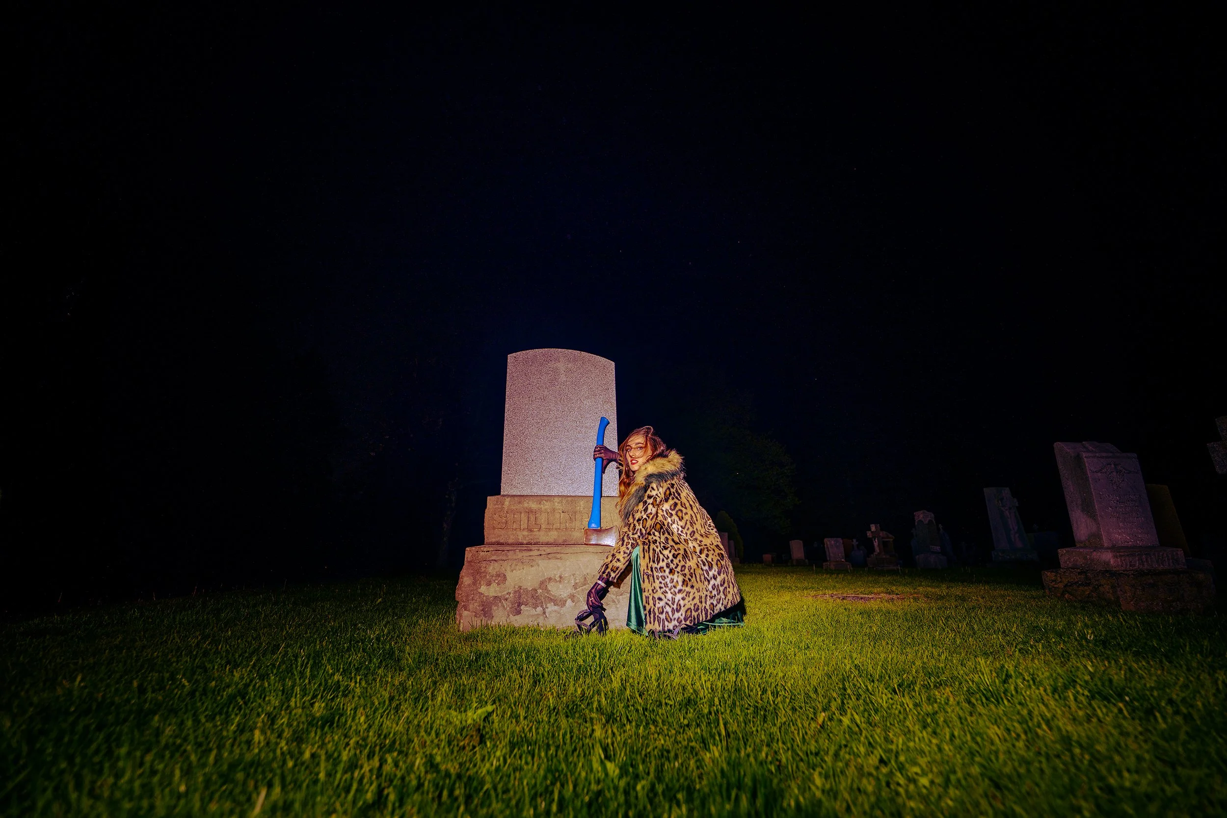 A woman with glasses and red hair, wearing a leopard print coat, crouches next to a tombstone at night in a graveyard. She holds a blue hookah pipe. The sky is dark, and there are other tombstones in the background.