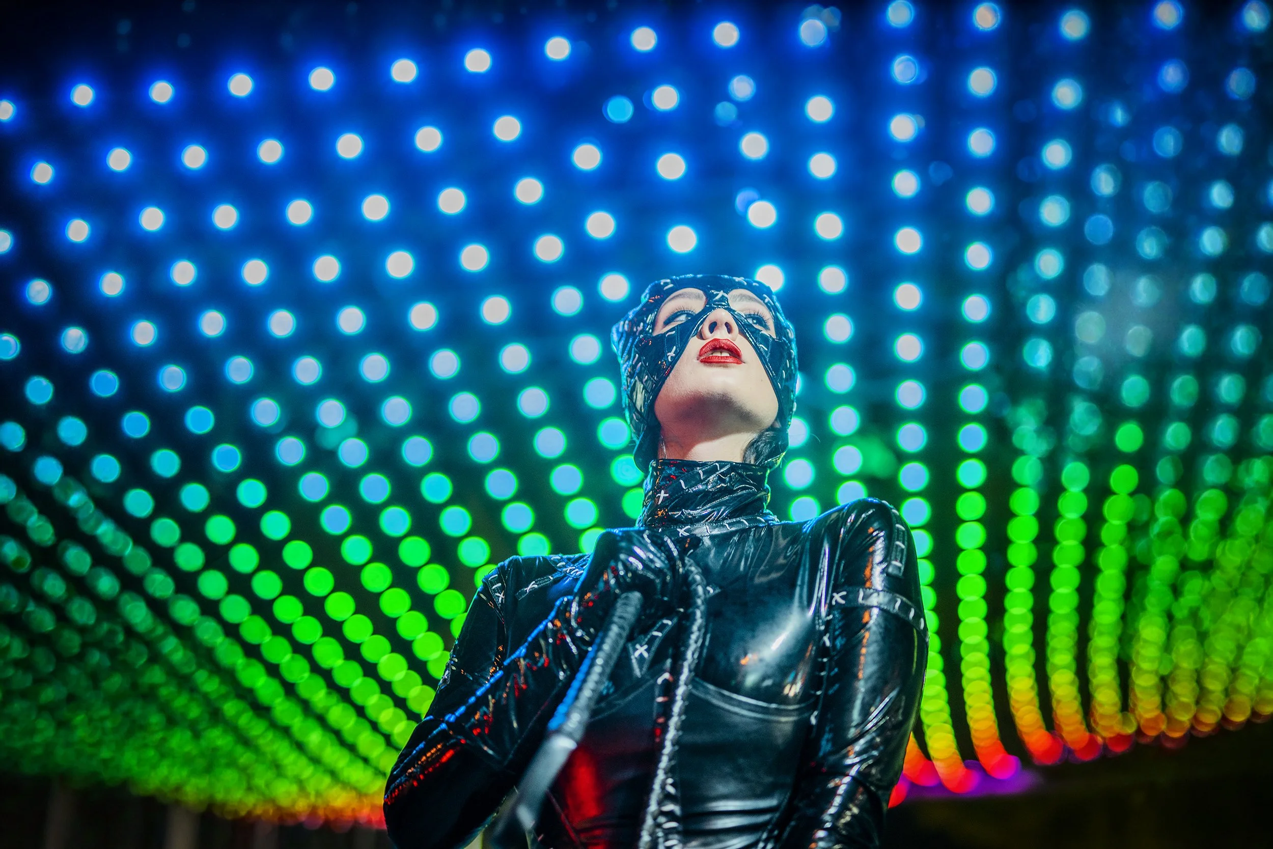 A person dressed in a shiny, black, futuristic outfit and headgear, standing in front of a colorful, illuminated LED backdrop with blue, green, yellow, and rainbow lights.
