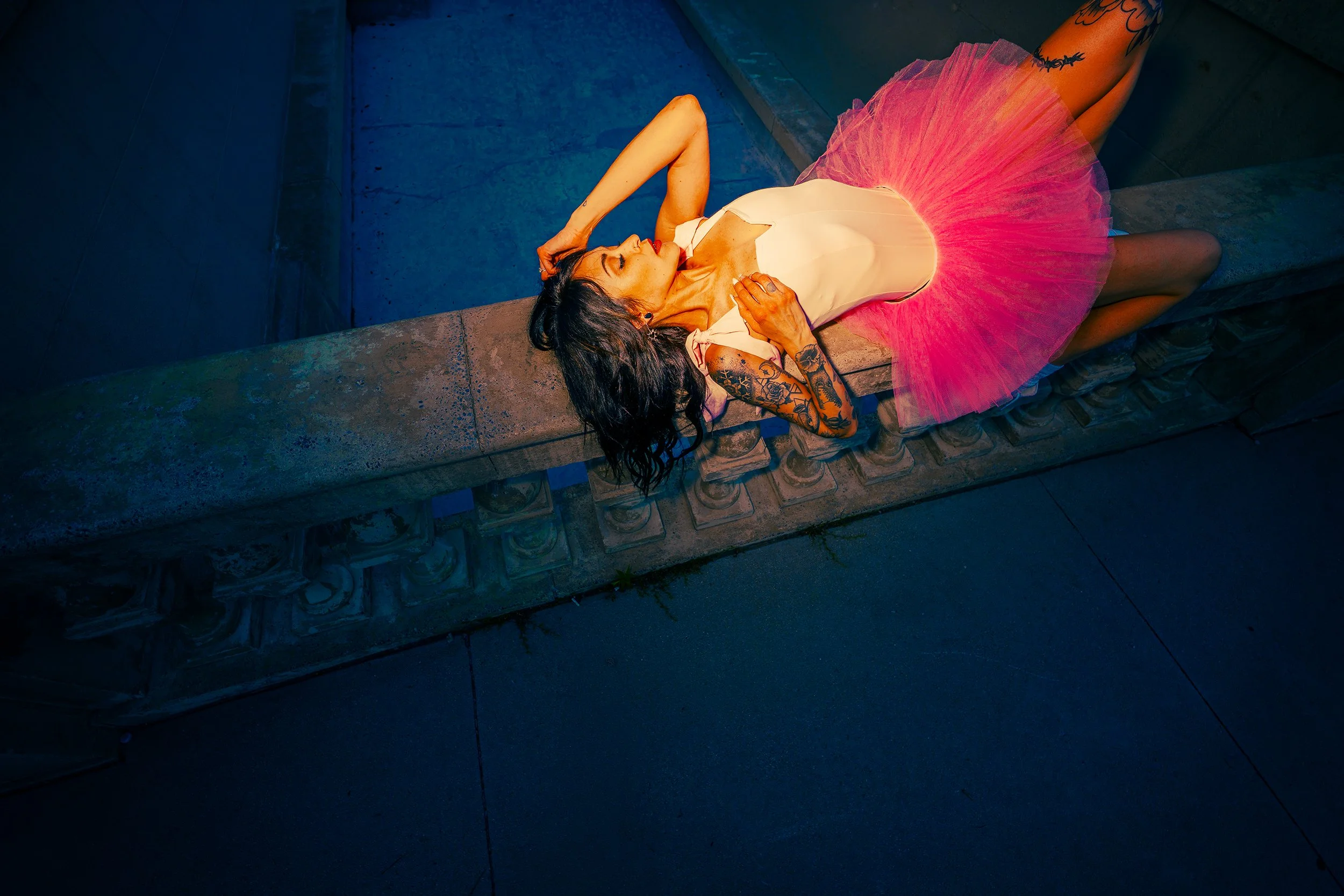 Woman lying on a stone ledge beside a pool, wearing a white dress with a pink tutu, with tattoos and dark hair, in an outdoor setting with blue lighting.