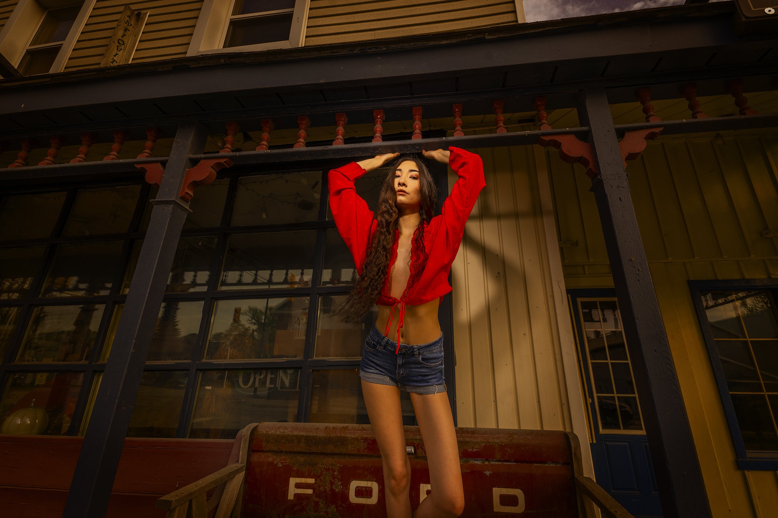 A young woman with long curly hair posing in front of a storefront with an "OPEN" sign. She is wearing a red top and denim shorts, standing with her hands on her head. The background includes wooden and glass elements of the building and a small wood