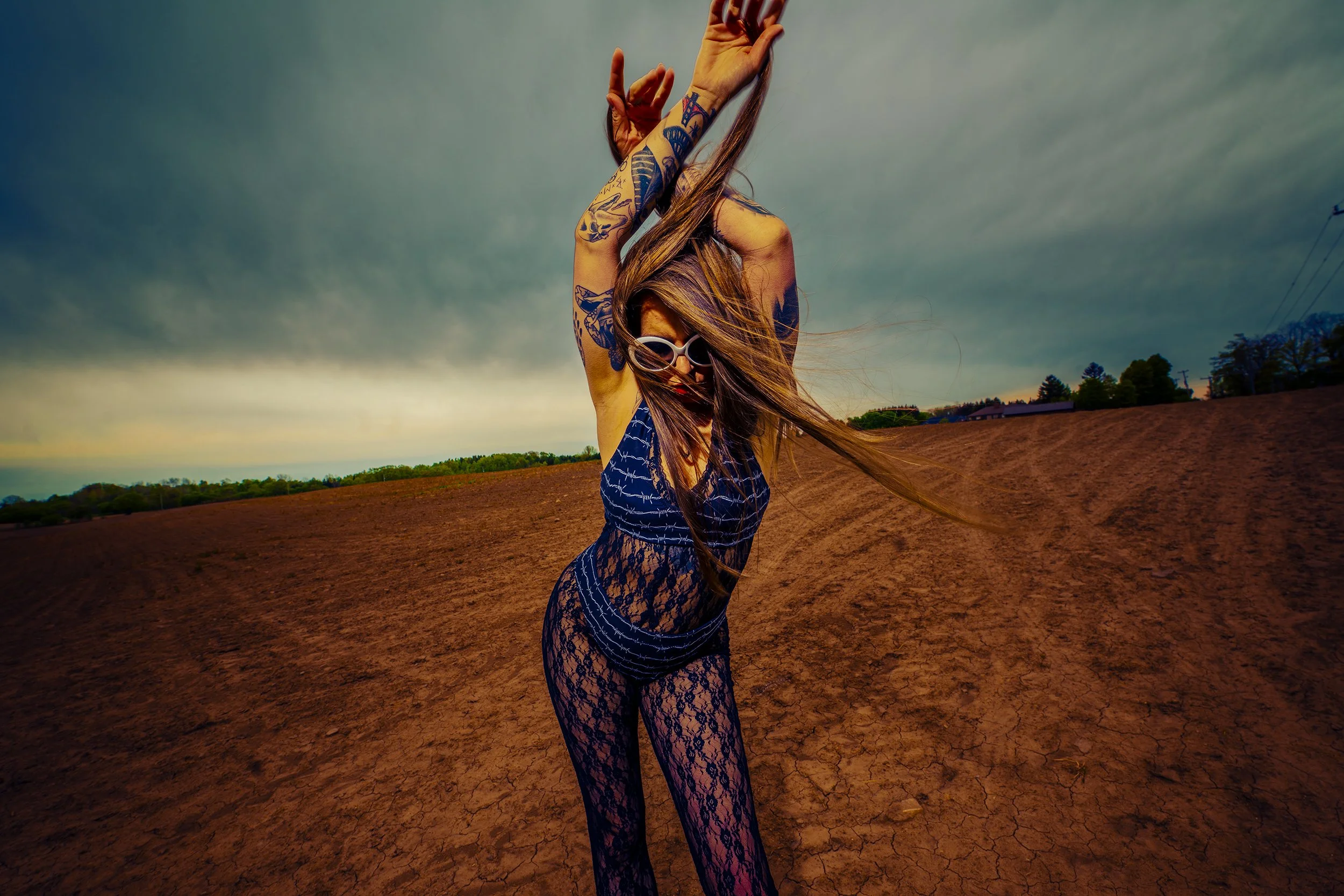A woman with long hair and tattoos, wearing sunglasses, a lace dress, and lace pants, standing on dry, cracked ground under a cloudy sky with her arms raised and flowing hair.