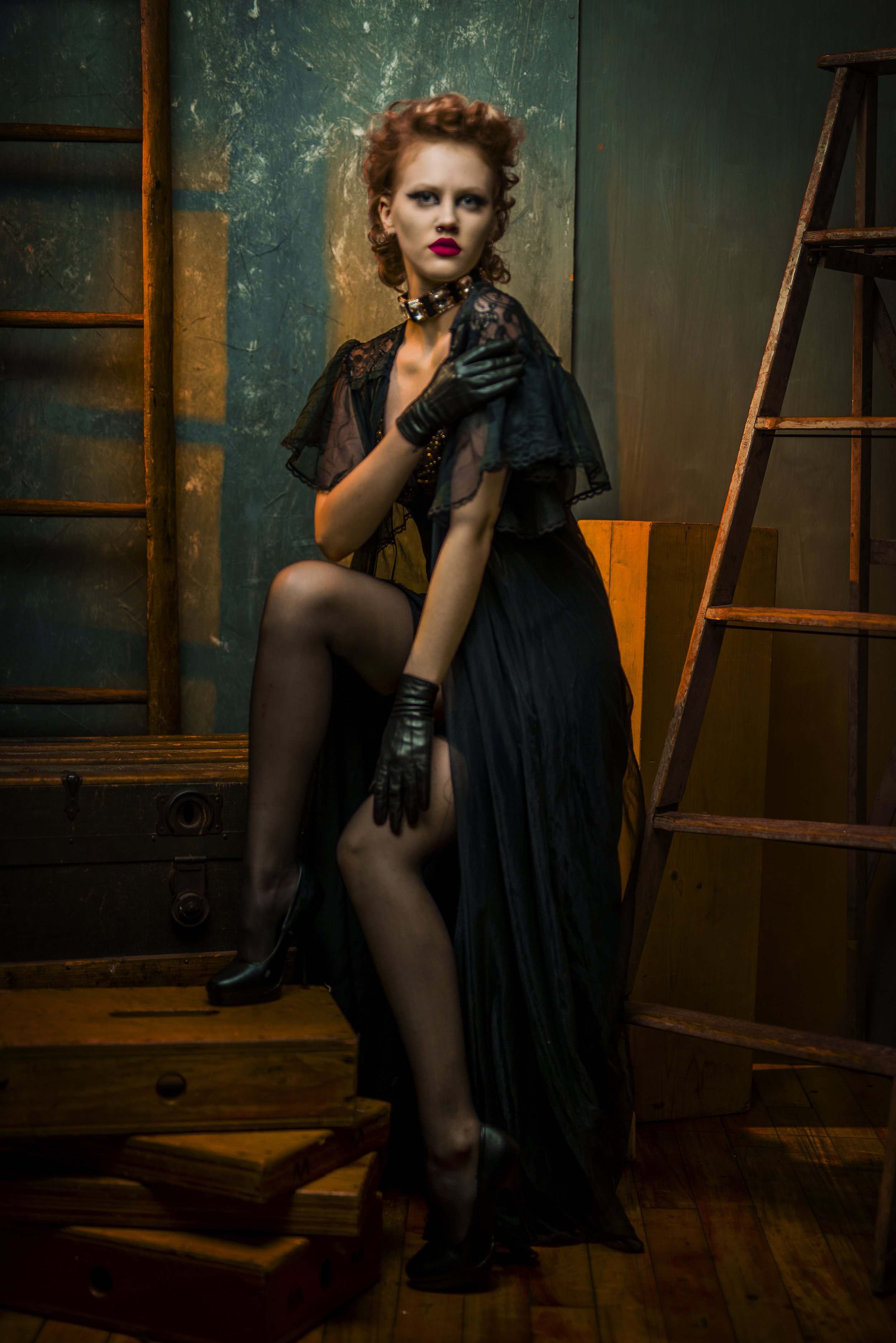 A woman with curly red hair, dark makeup, and red lipstick is sitting on wooden stairs, wearing a black dress with sheer lace sleeves, black gloves, and thigh-high stockings. She is touching her neck with one hand and has her other hand on her thigh,