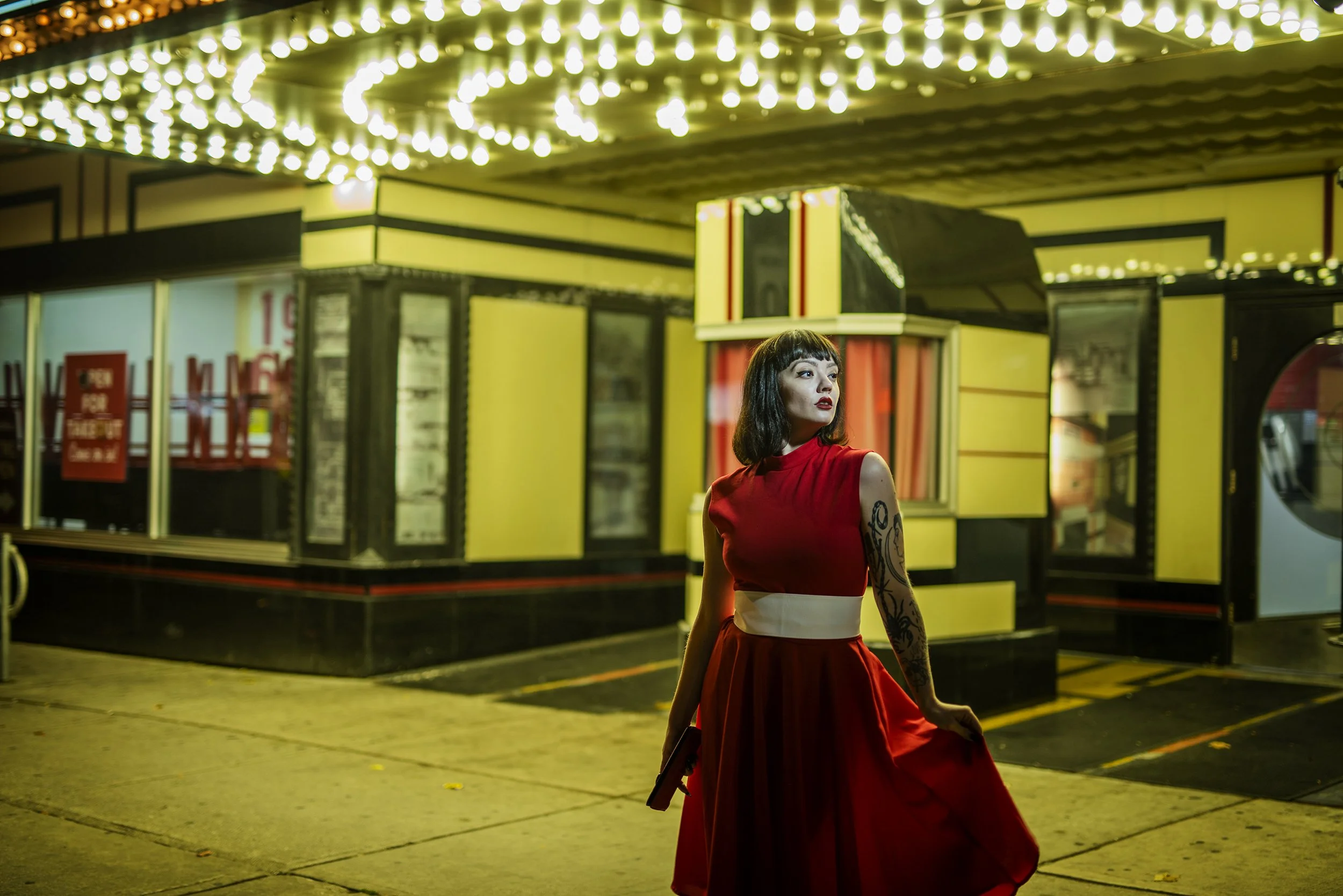 A woman in a red dress standing outside a brightly lit carnival at night. The background features a yellow and black snack booth with string lights overhead.