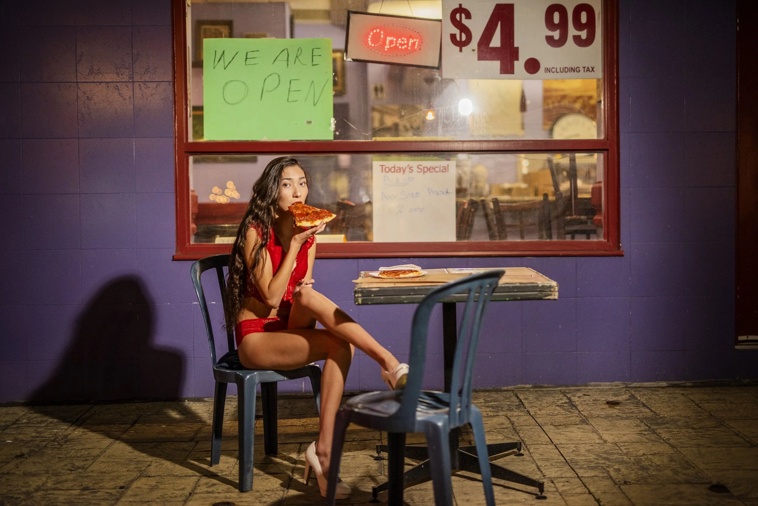 A woman in a red dress sitting outside a pizza restaurant at night, eating a slice of pizza. The restaurant has signs indicating it is open, with one sign in the window saying $4.99 including tax, and another advertising a special deal for an extra l