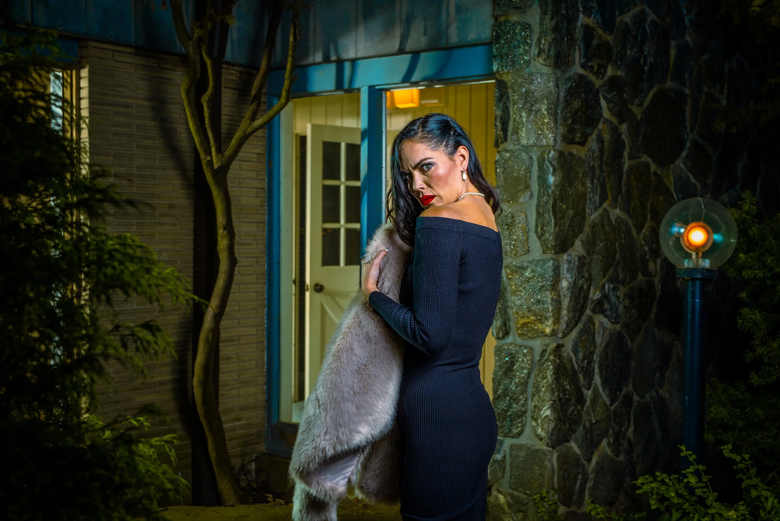 A woman with dark hair and red lipstick holding a dog in front of a house at night with colorful lighting.