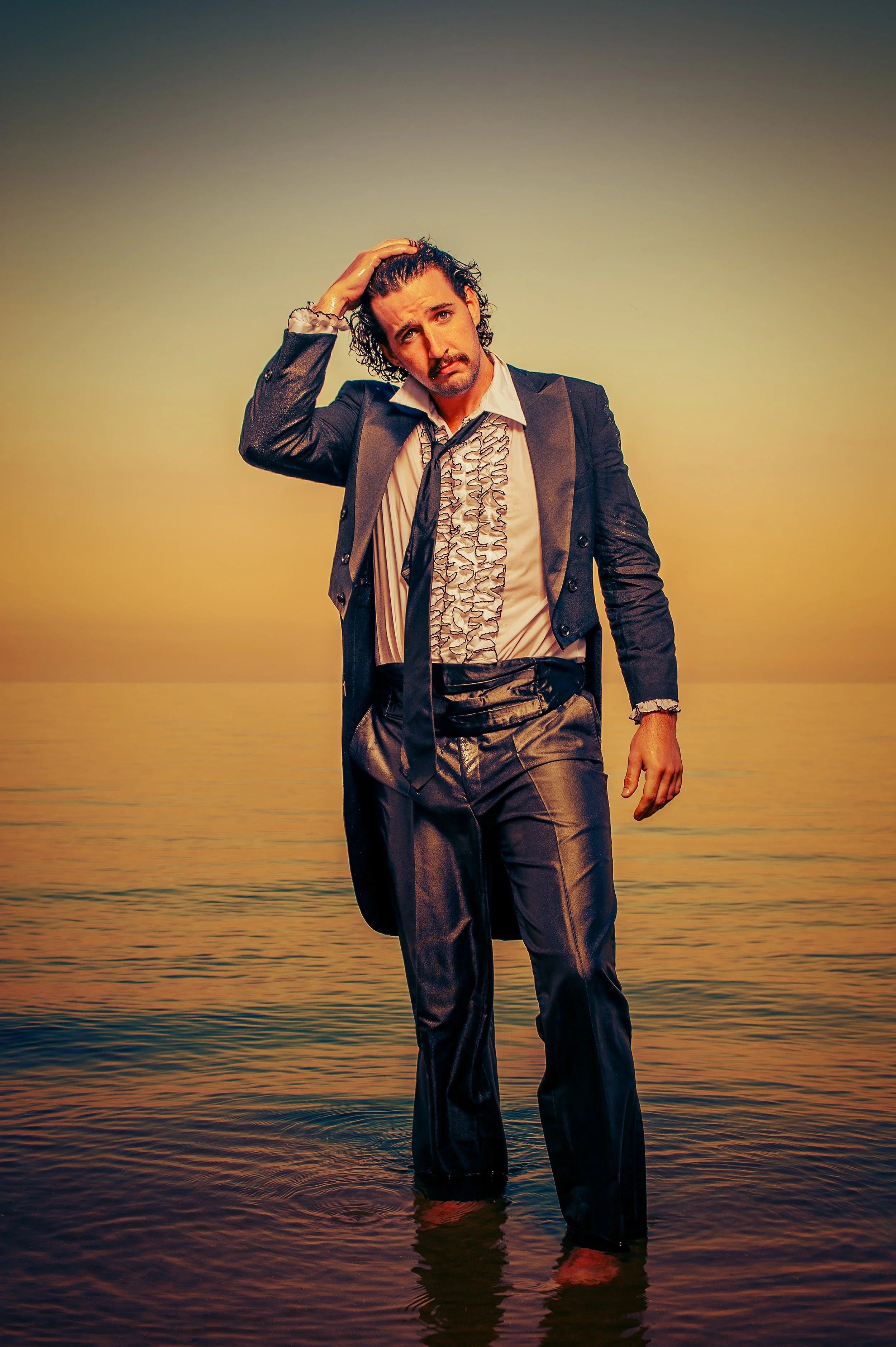 A man with wet hair and a mustache dressed in a tuxedo with ruffled shirt, standing in shallow water at sunset, touching his head with a contemplative expression.