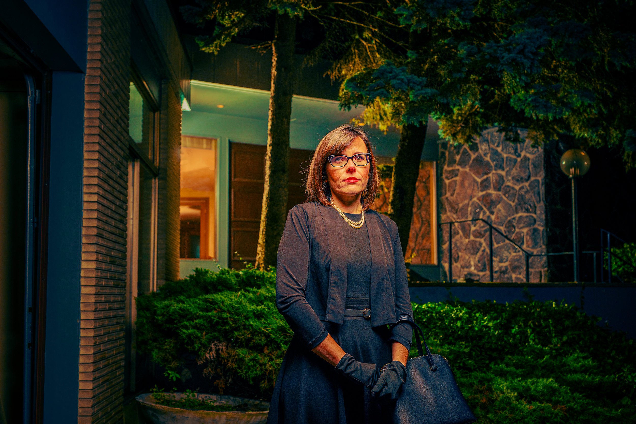 A woman standing outside at night in front of a modern building with brick and stone walls, surrounded by trees and greenery, dressed in a dark dress with pearl necklace, wearing glasses, and holding a black handbag.