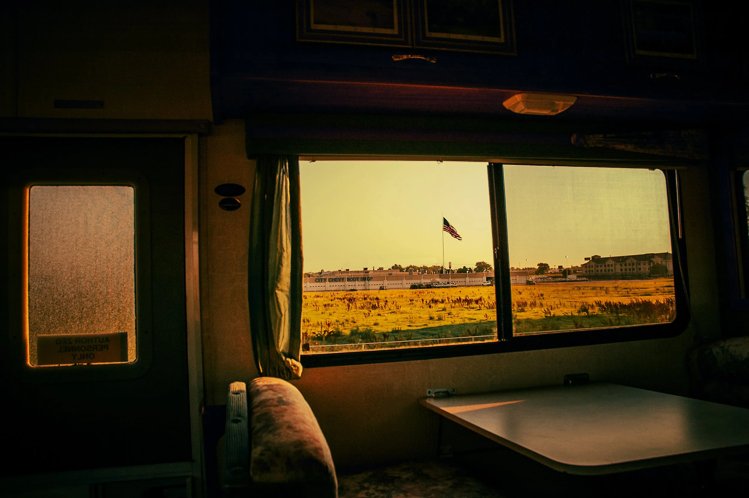 Interior of a vehicle with a window showing a sunset sky, a field, and buildings including a sign that reads 'City Chevy Body Shop.'