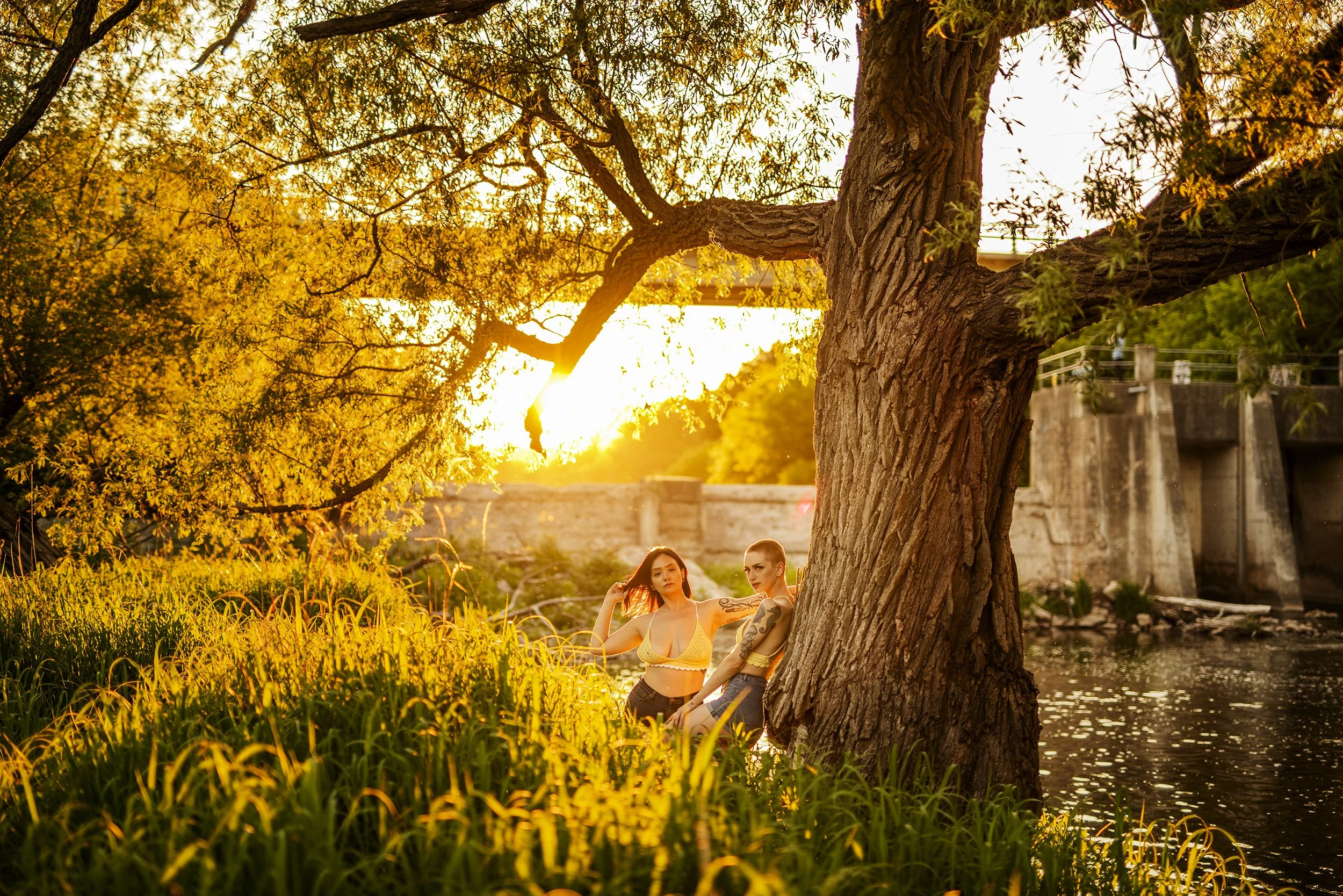 Two women in yellow tops and shorts stand by a riverbank under a large tree at sunset with the sun setting behind them.