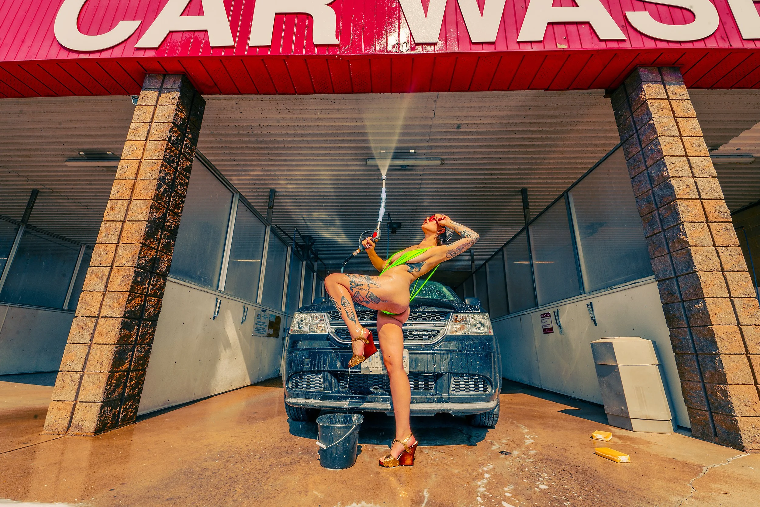 A person with tattoos wearing a bright green swimsuit and high heels is dancing and posing while washing a black car at a car wash station with a roof and brick columns, under a pink sign that reads 'CAR WASH.'