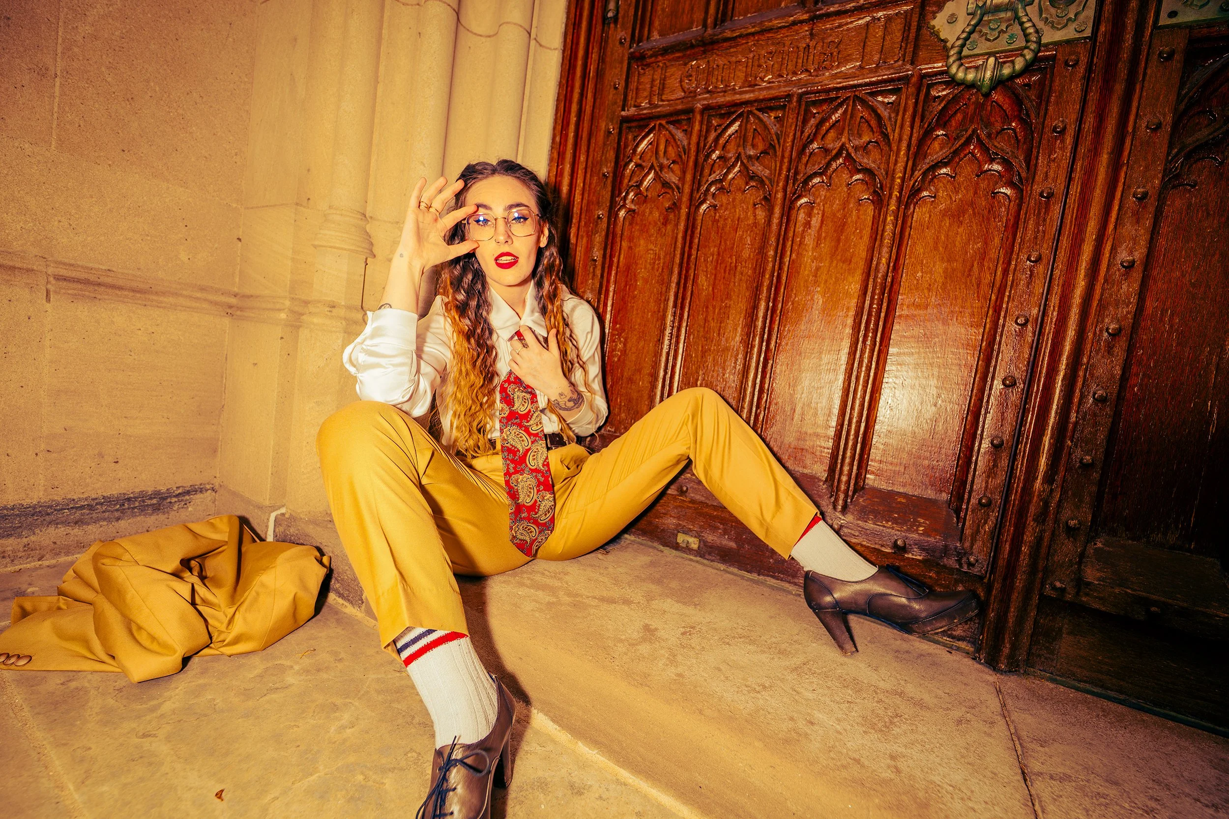 A woman with glasses, curly hair, and red lipstick sitting on the floor next to a large wooden door. She is dressed in yellow pants, a white shirt, and a red patterned tie, with a yellow jacket on the floor beside her. She is holding her glasses with