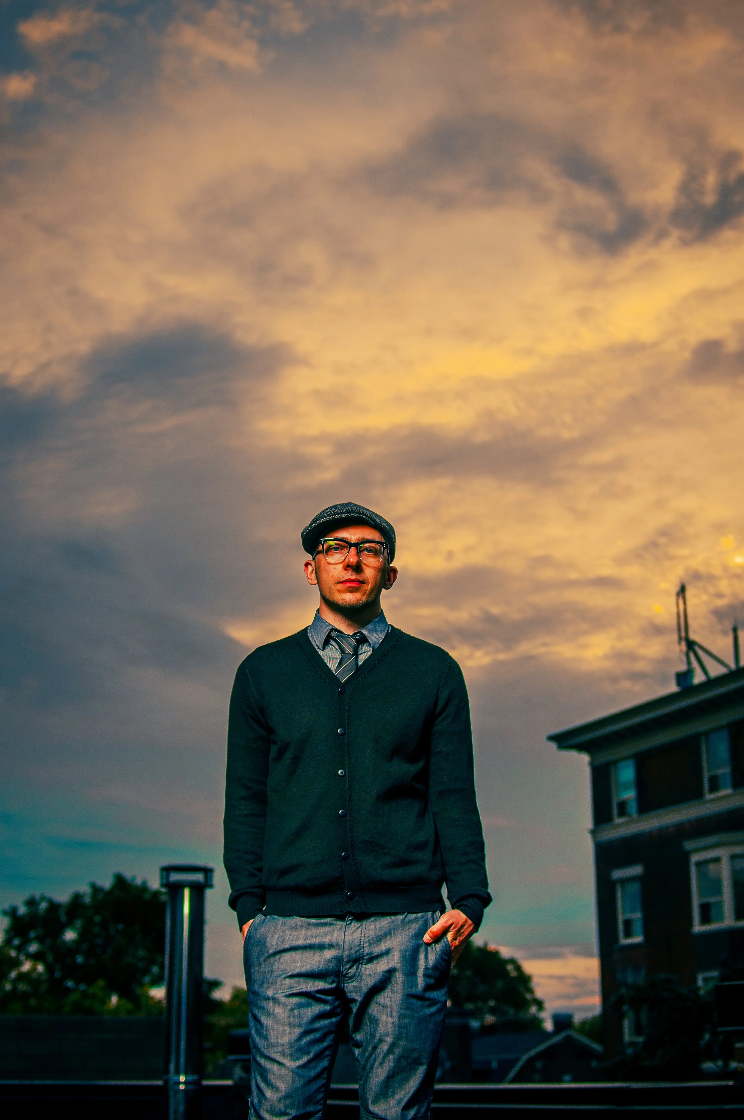 A man dressed in a black sweater and gray pants standing outdoors on a rooftop during sunset, with a cloudy sky and buildings in the background.