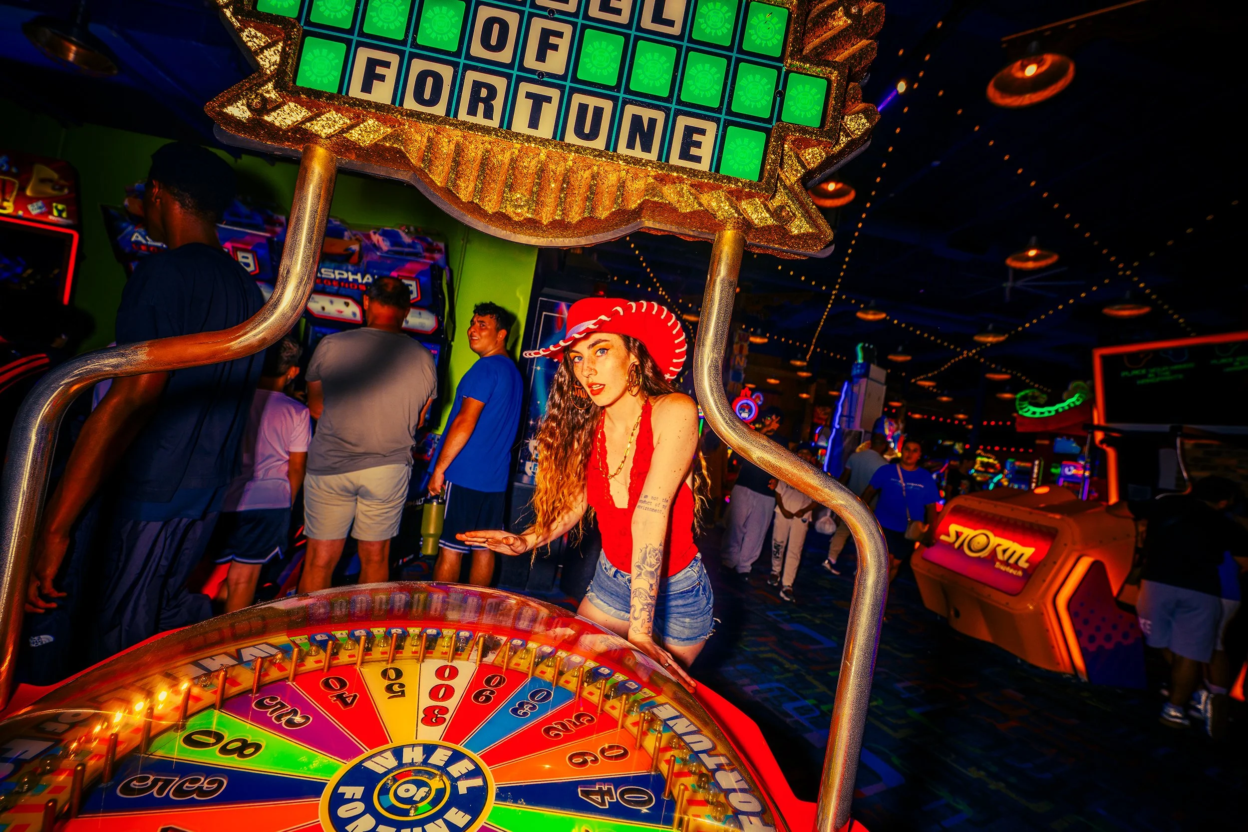A young woman with long curly hair, tattoos, and wearing a red dress and red cowboy hat, standing near a colorful spinning wheel at an arcade or amusement venue. Behind her, a group of people are seen in a dimly lit arcade with various game machines 