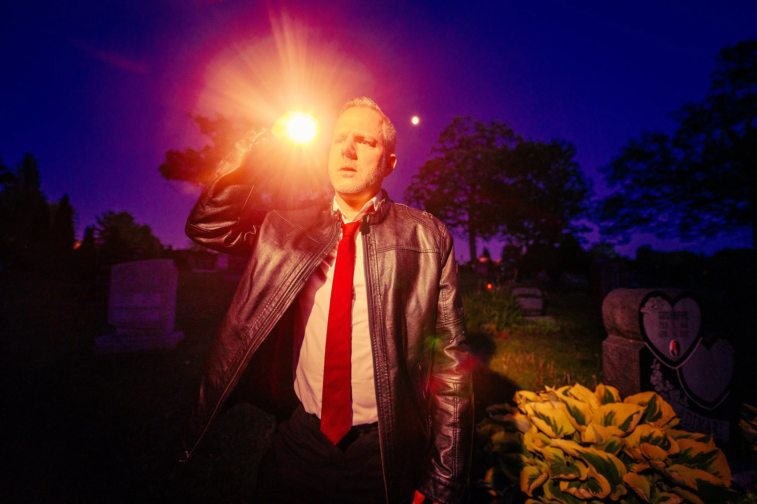 A man in a leather jacket and red tie stands outdoors at night, holding a lit flare to his forehead, with a purple sky, trees, gravestones, and flowers in the background.