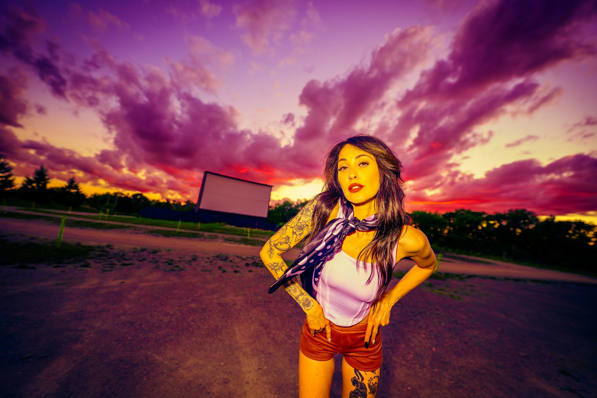 A woman with tattoos on her arms and leg, wearing a white tank top, reddish-brown shorts, and a purple polka dot scarf, standing outdoors during a vibrant sunset with colorful pink and purple clouds in the sky.